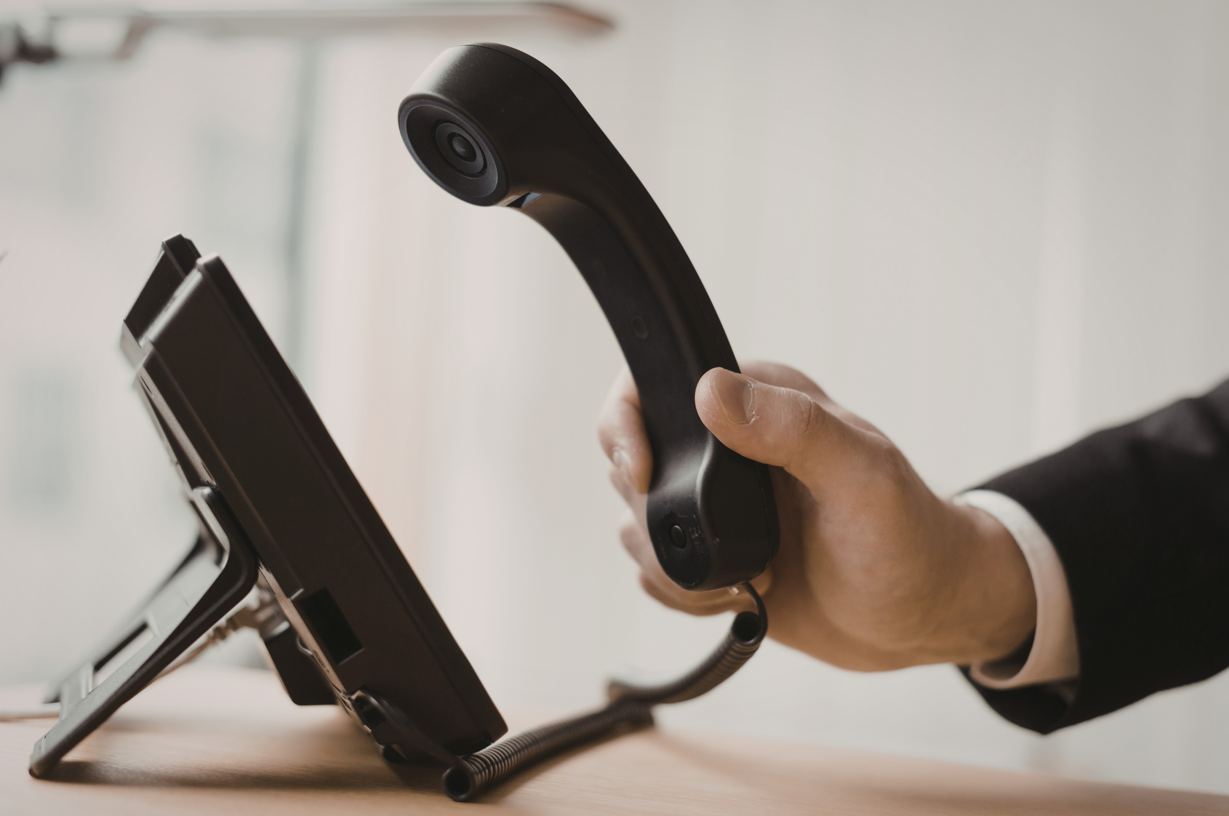 A person in business attire holding a black desk phone handset, with the phone placed on a stand on a wooden surface.
