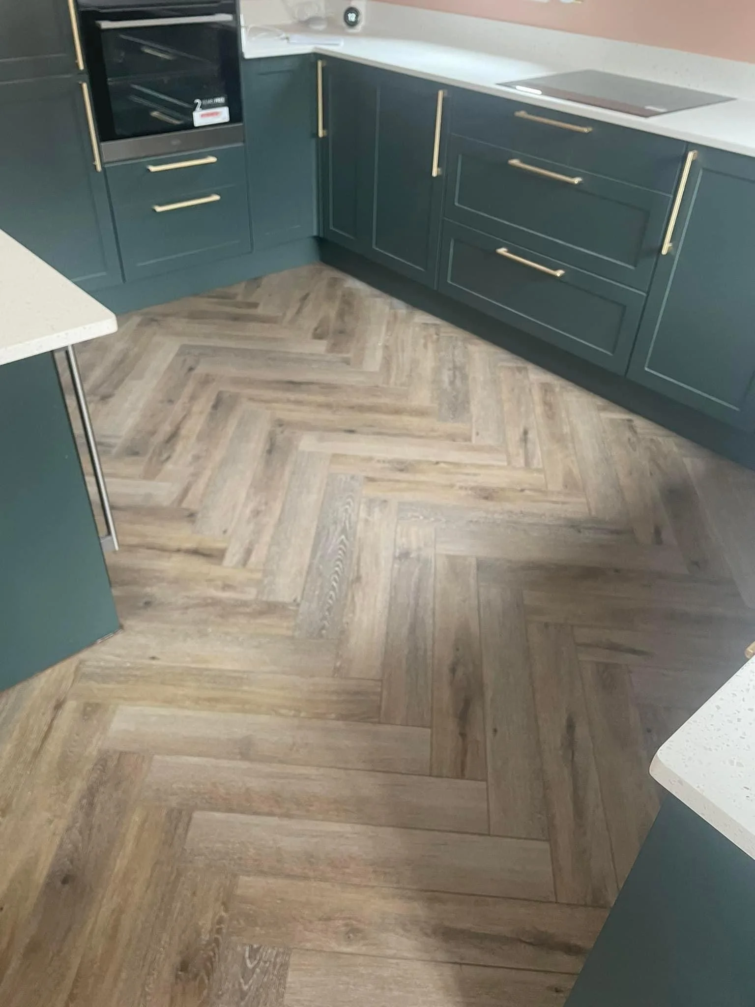 Modern kitchen with herringbone wood floor, dark green cabinets, gold handles, white countertops, and integrated oven.