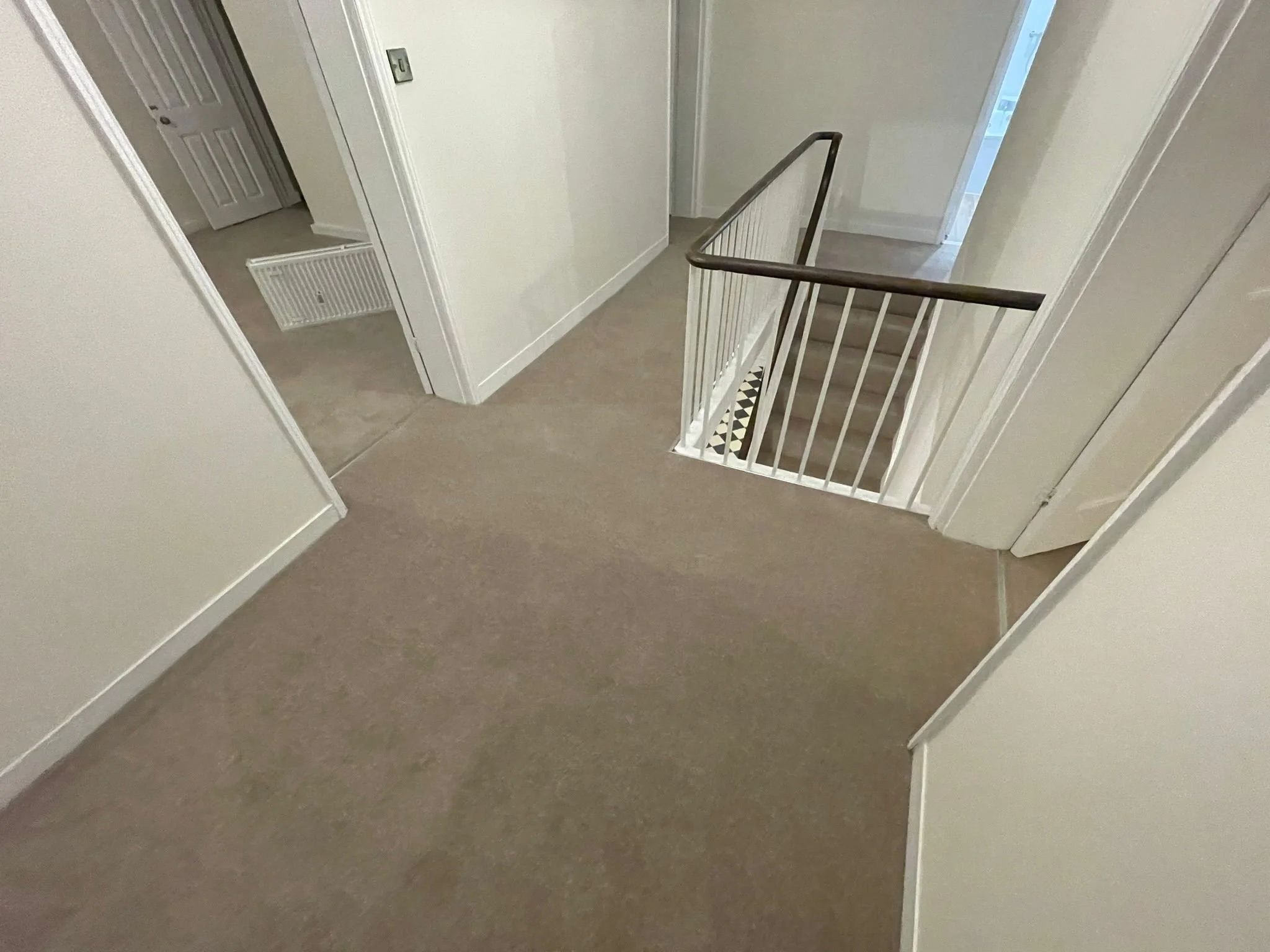 Carpeted hallway in a residential home with plain white walls, a staircase with a white railing, and a doorway leading to another room.