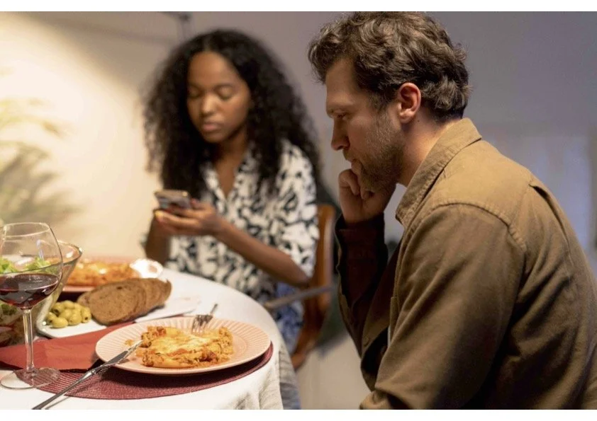 A young couple eating dinner in silence, the man looking pensive and vacant as the woman taps away at her phone