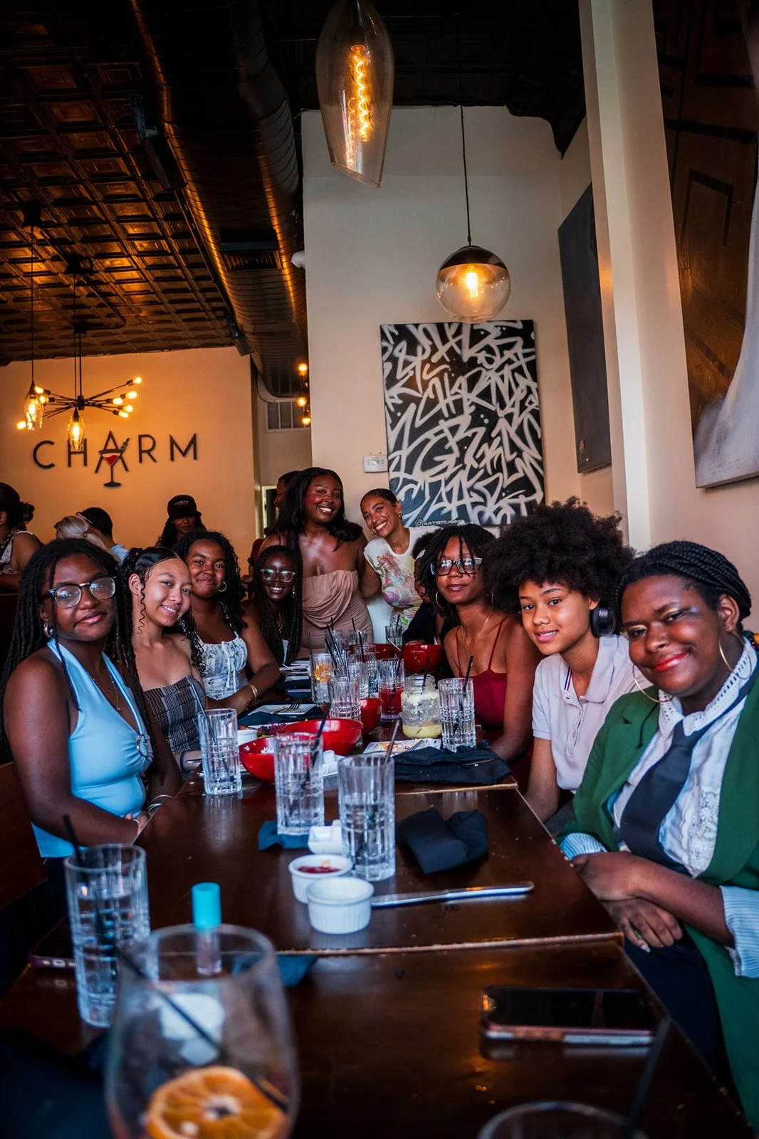 A group of women and girls gathered around a restaurant table, smiling for the photo, with drinks and bowls on the table. The restaurant decor includes abstract artwork and hanging light fixtures, with a sign on the wall that reads 'CHARM'.