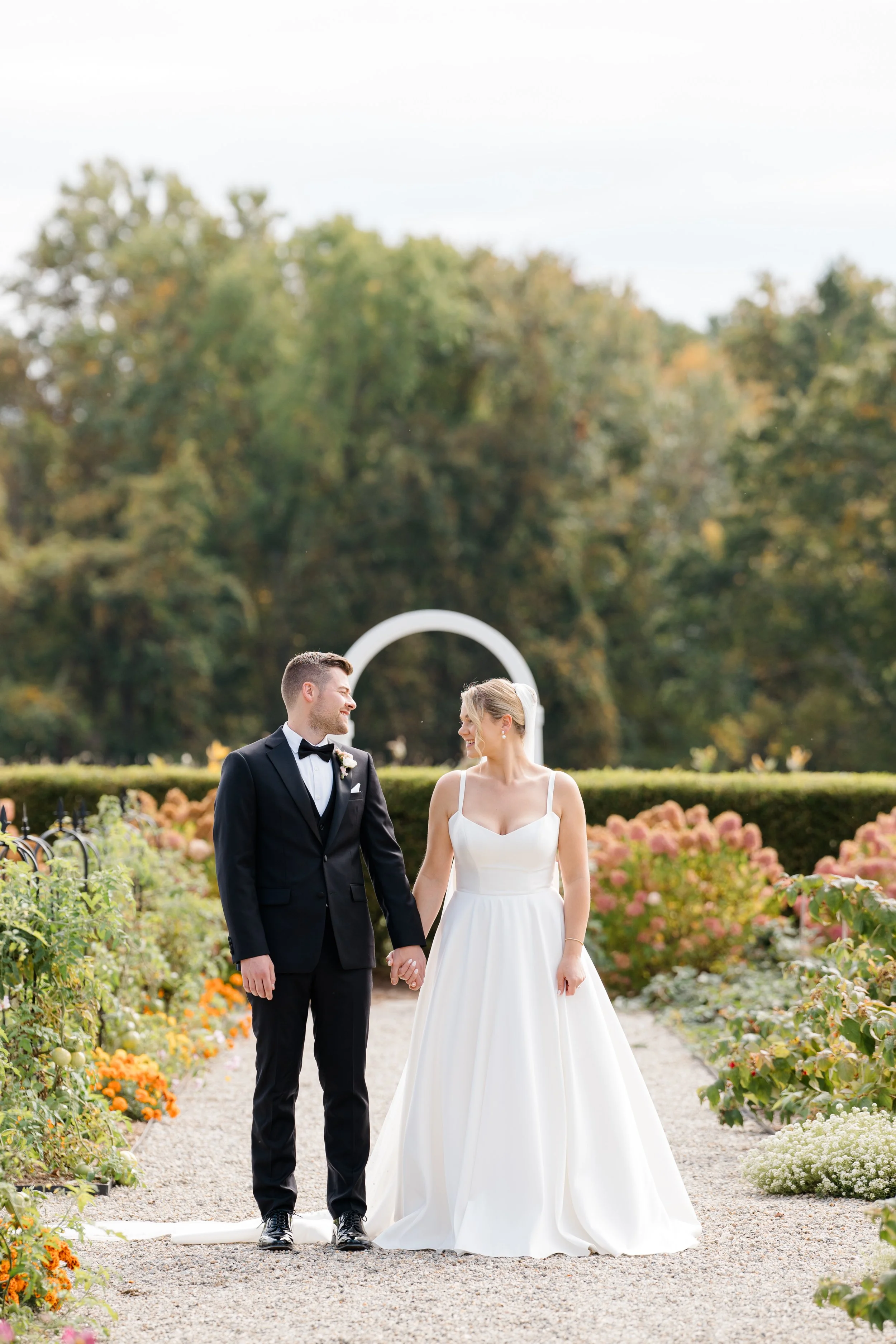 A bride and groom holding hands, walking on a garden path during their wedding, with colorful flowers and trees in the background.