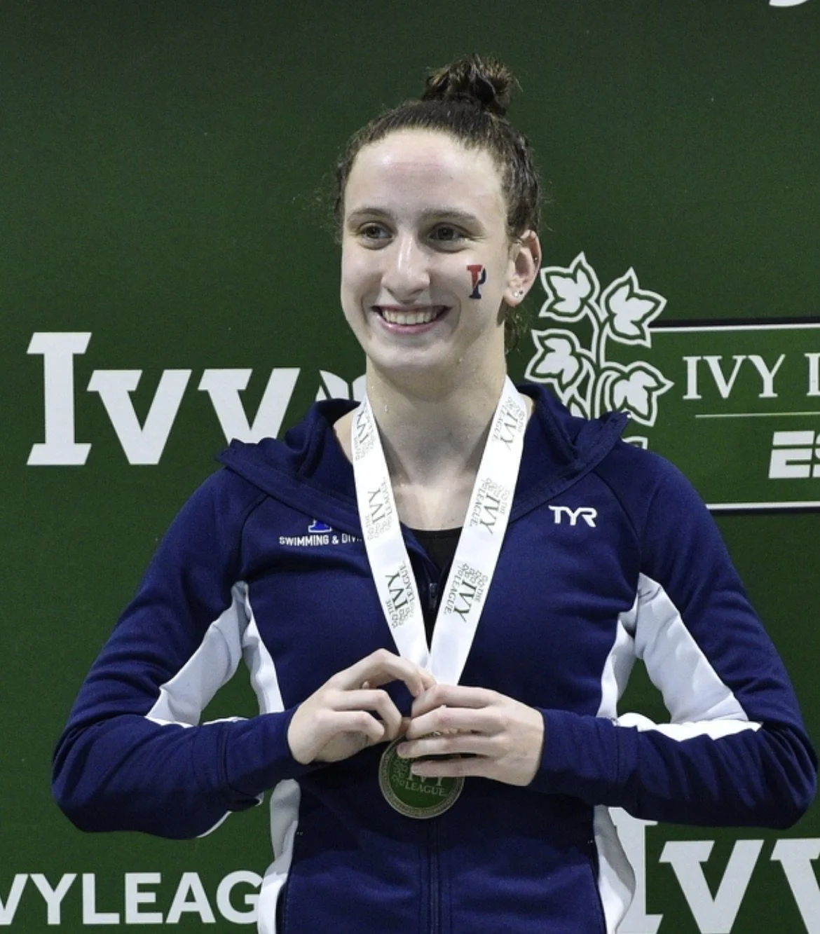 Person wearing medal and dark blue athletic jacket in front of Ivy League backdrop at the 2019 Ivy League swimming & diving championships.