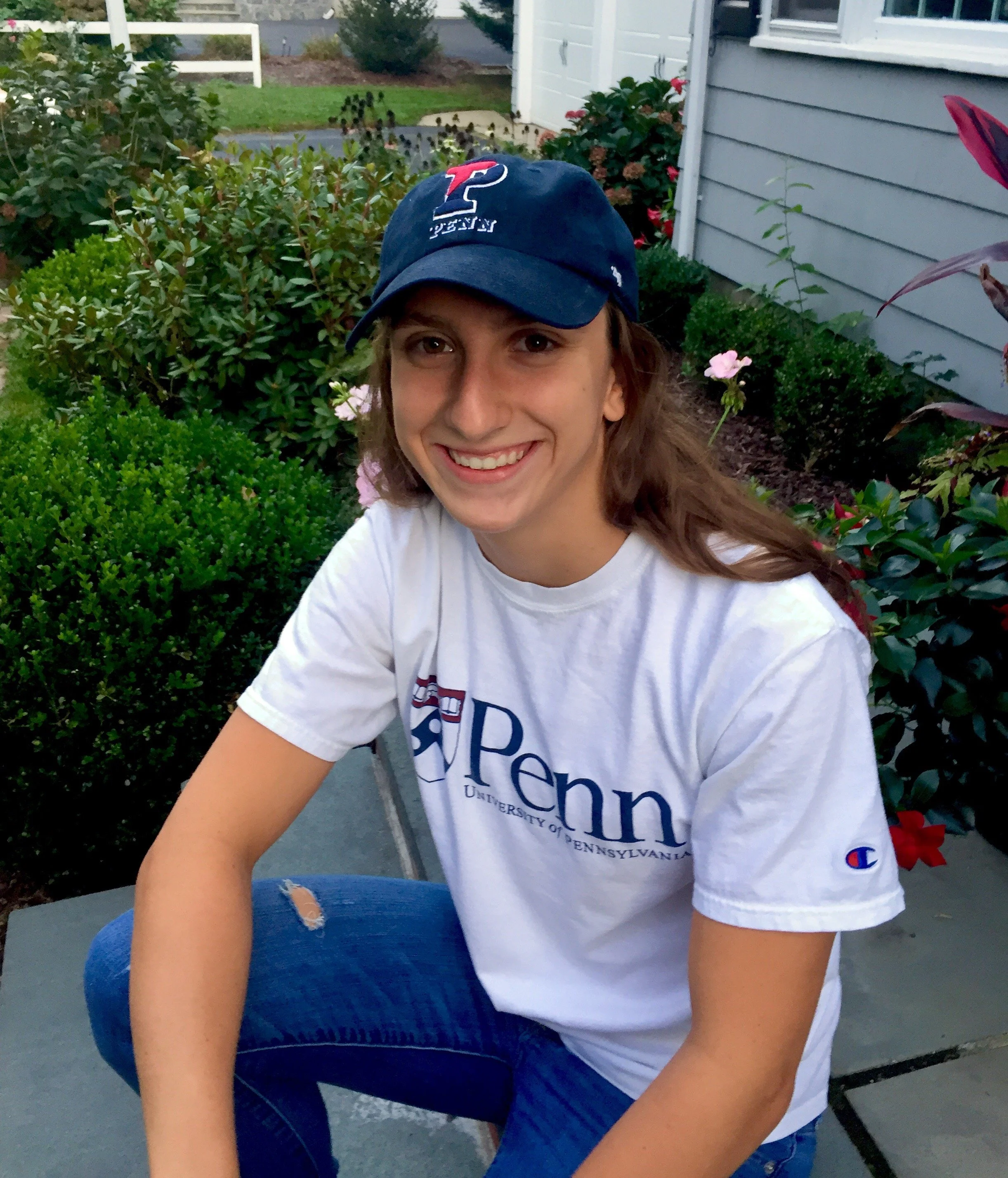 A person wearing a University of Pennsylvania t-shirt and cap, sitting on a stone path, surrounded by greenery and flowers.