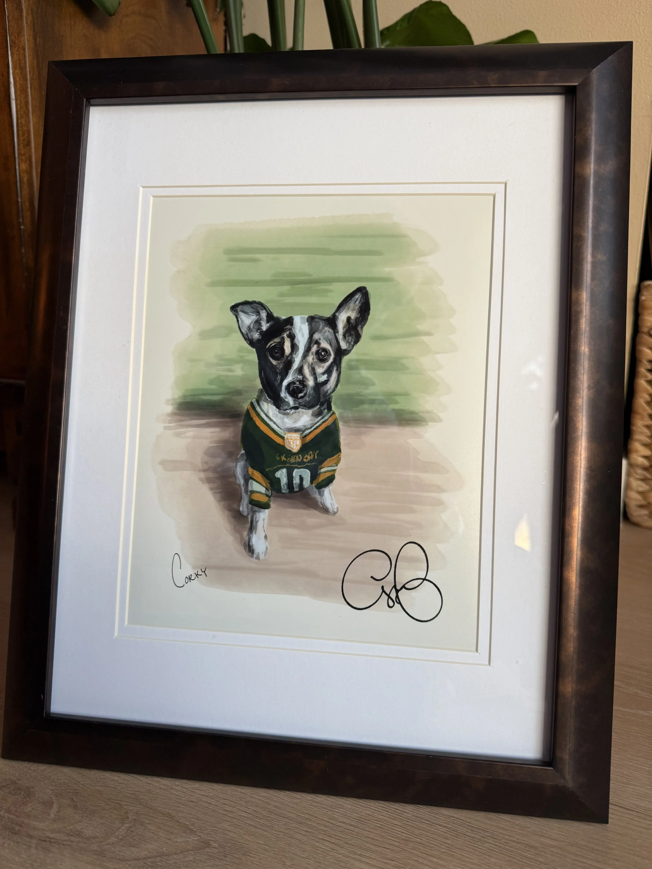 A framed watercolor portrait of a small dog wearing a green sports jersey. The dog has black, white, and gray fur with large ears and expressive eyes. The background has green and beige watercolor strokes. The frame is black with a white mat, and the