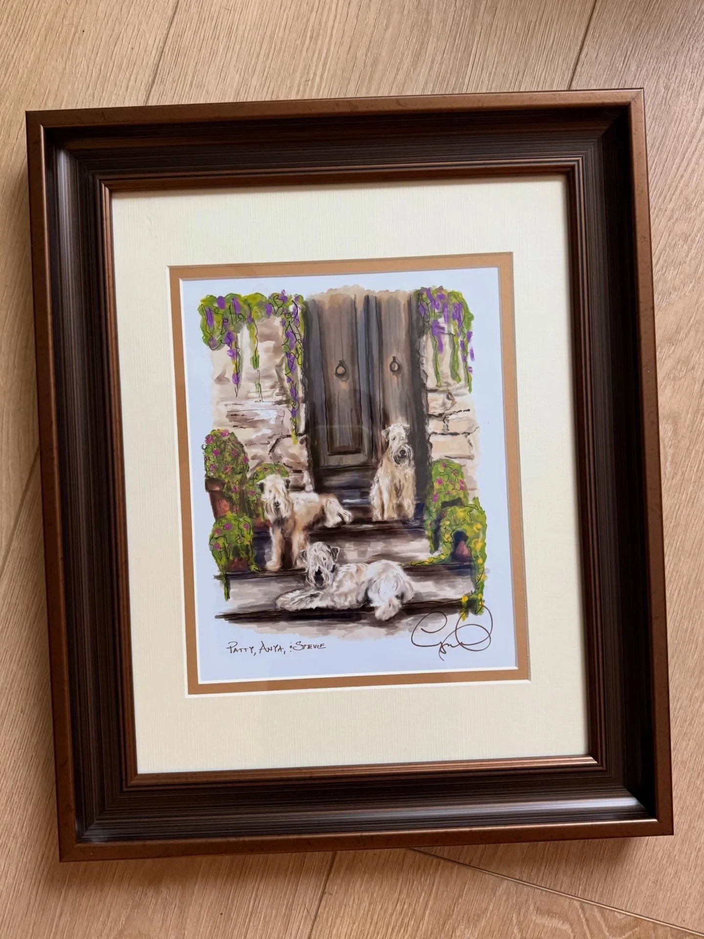 Framed watercolor painting of three golden retrievers sitting and lying in front of a stone wall with a door, surrounded by greenery and hanging flowers.
