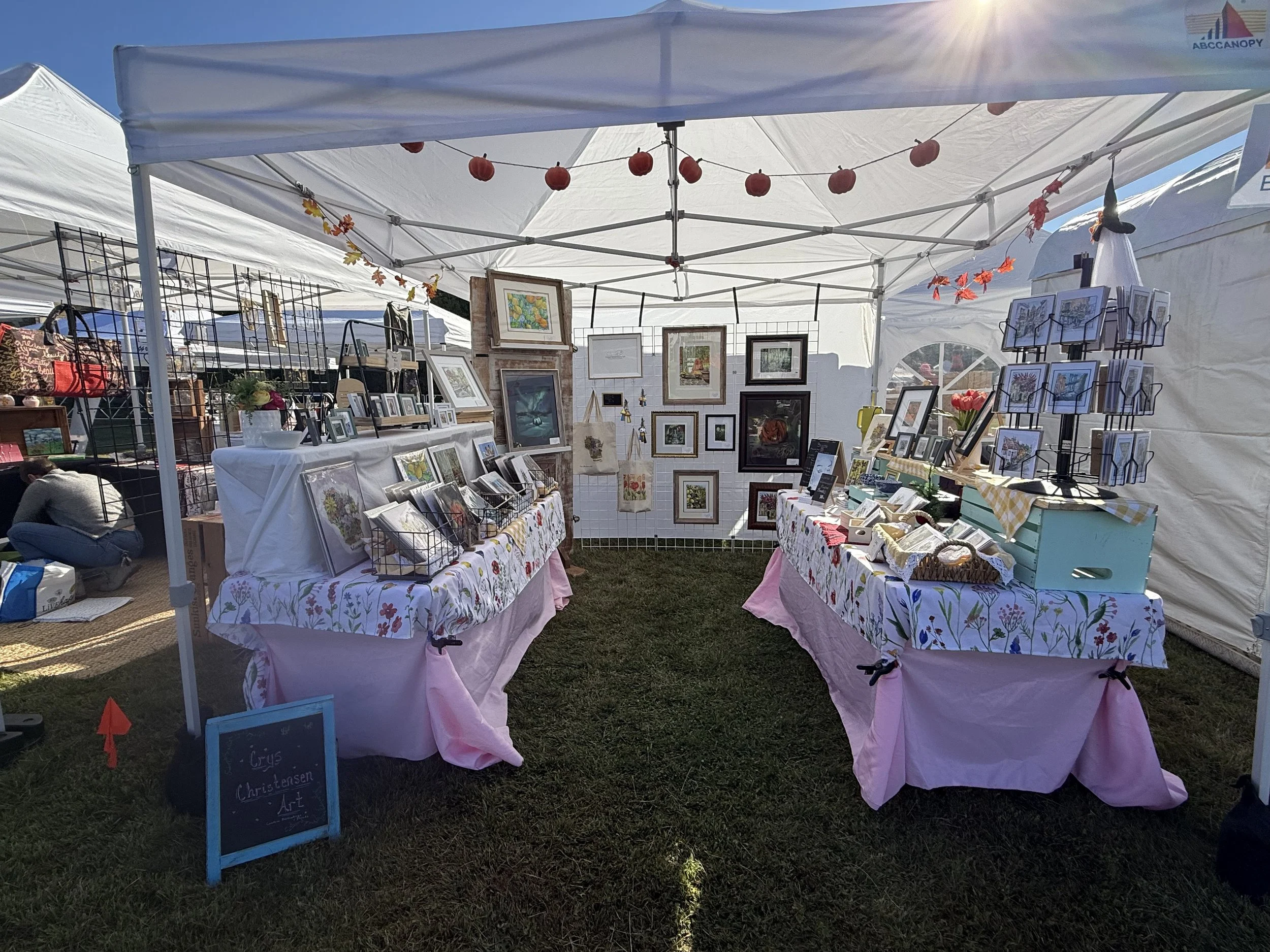 Outdoor art fair booth with framed artwork on display, decorated with string lights, paper lanterns, and autumn leaves, under a white canopy on a sunny day.