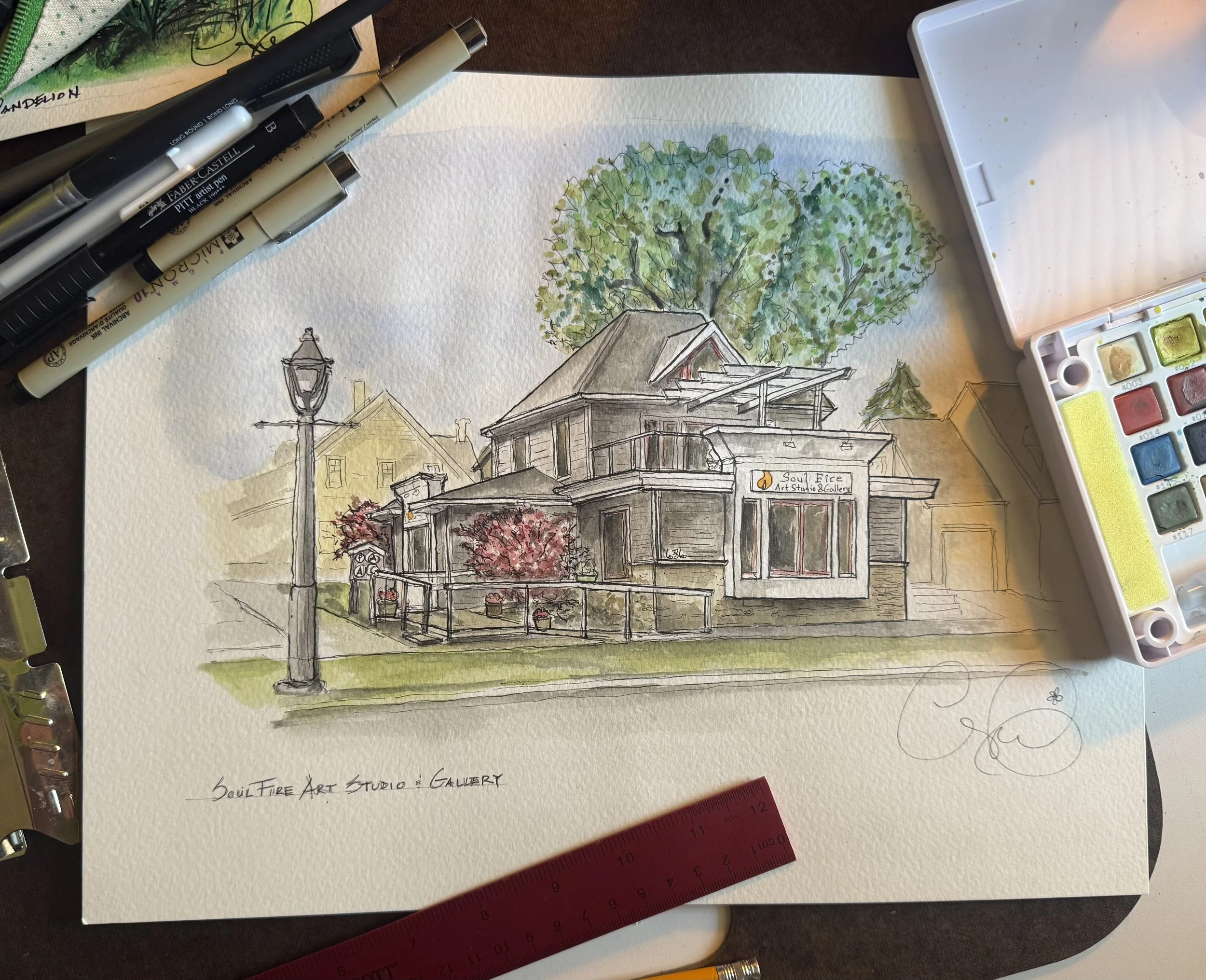 A watercolor and ink drawing of a house with a porch, trees, and a lamp post, with art supplies, including pens, watercolor paints, and a ruler, around the artwork.