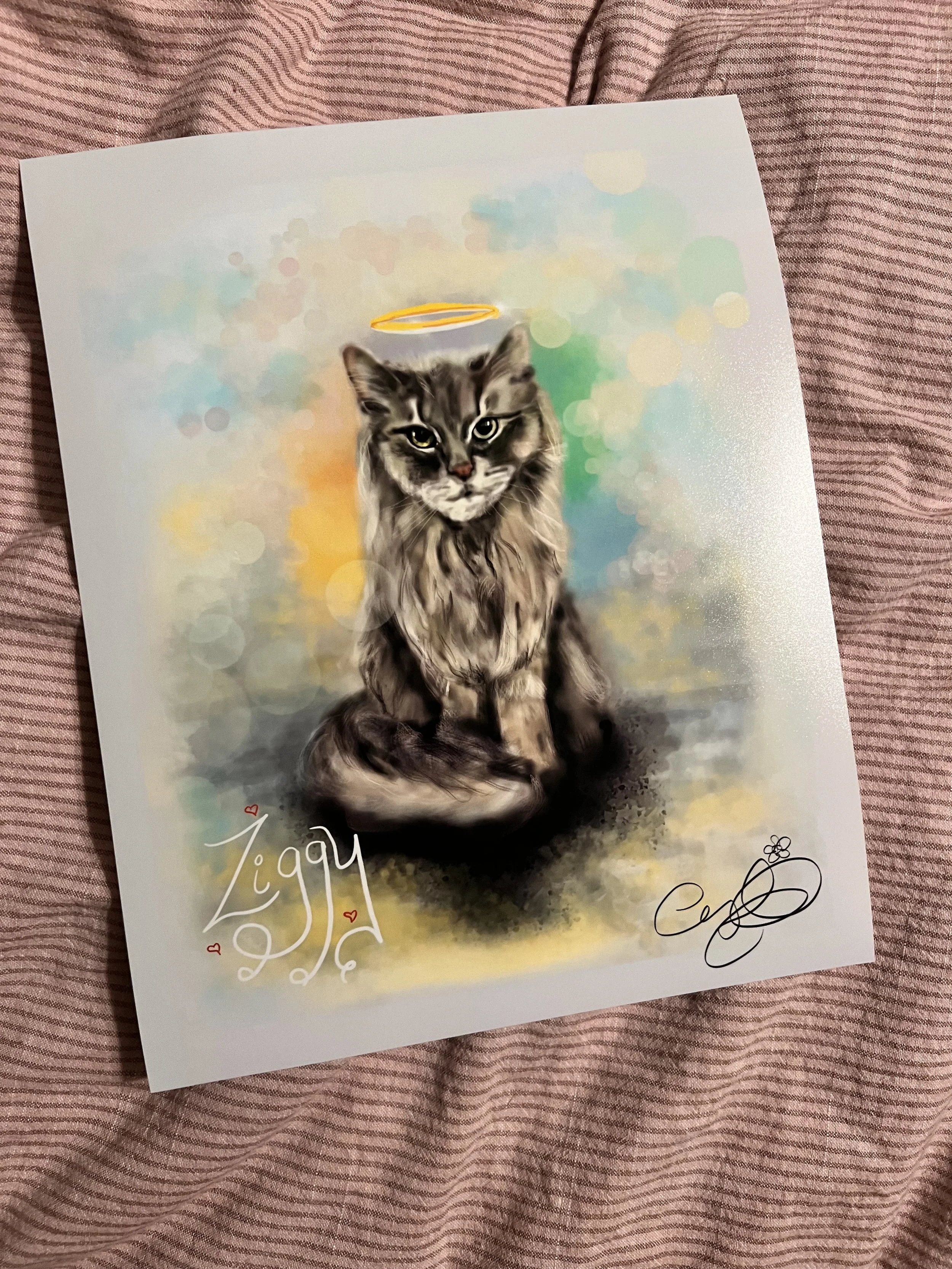 A watercolor painting of a gray tabby cat with a halo above its head, sitting on a surface with a colorful, blurred abstract background. There are handwritten signatures and small red hearts on the artwork.