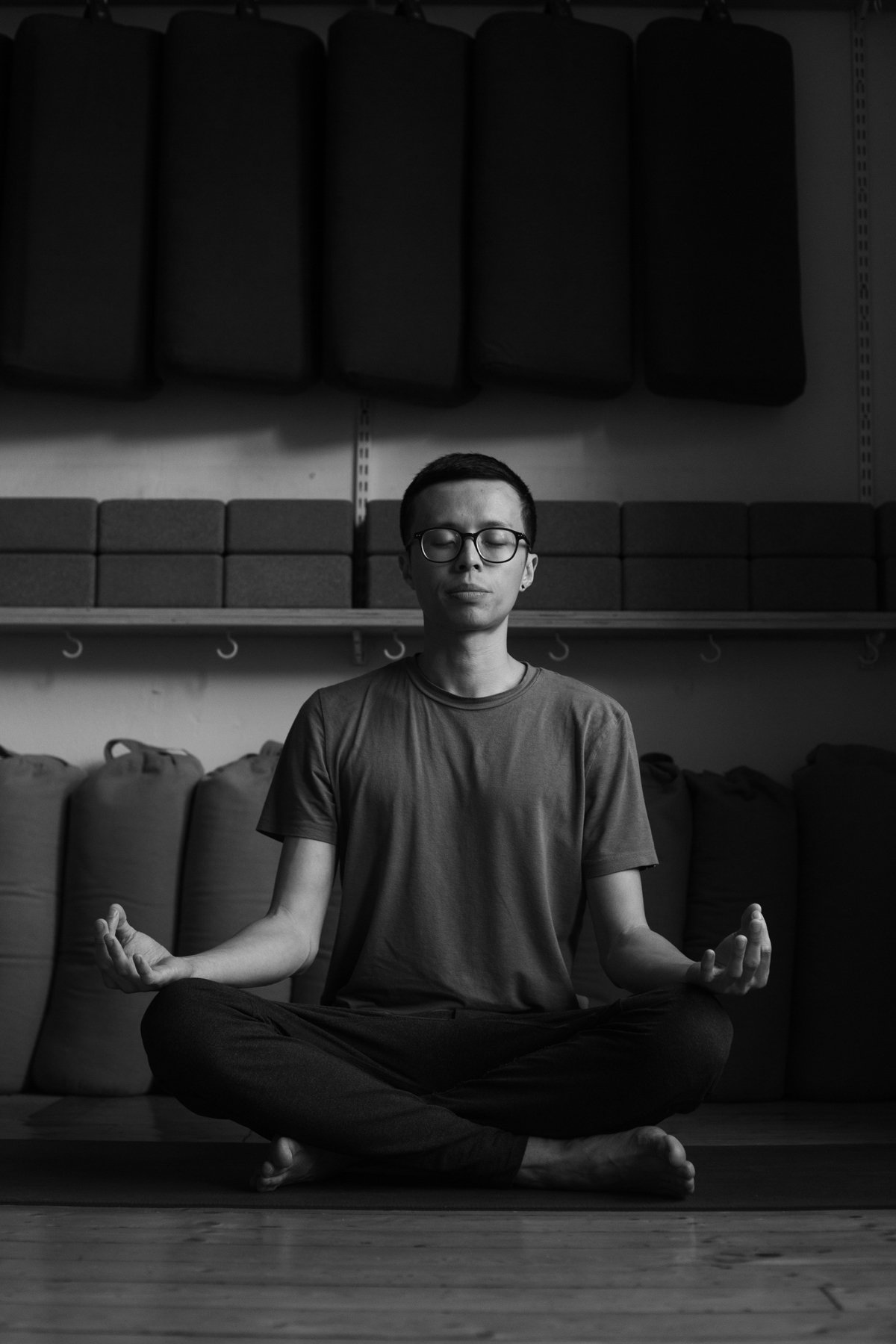 A young man wearing glasses sits cross-legged on the floor in a meditation pose with his eyes closed, in a room with foam padding and bags in the background.