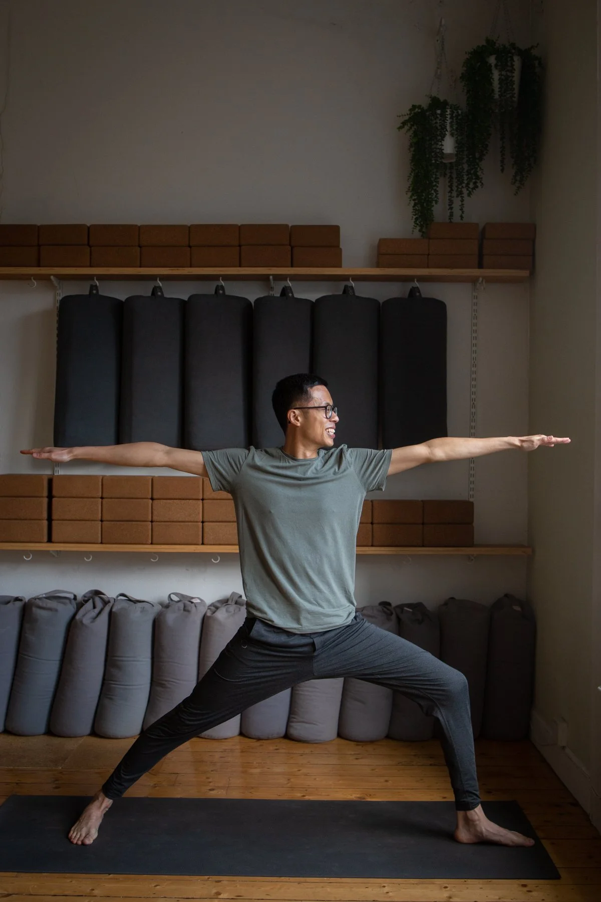 A man doing yoga in a lunging pose indoors with shelves, cushions, and potted plants in the background.