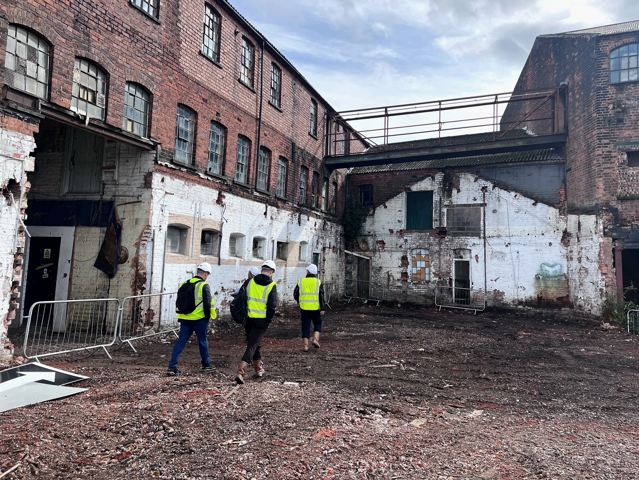 Three construction workers wearing yellow safety vests and white helmets walking through a vacant construction site with old brick buildings and debris.