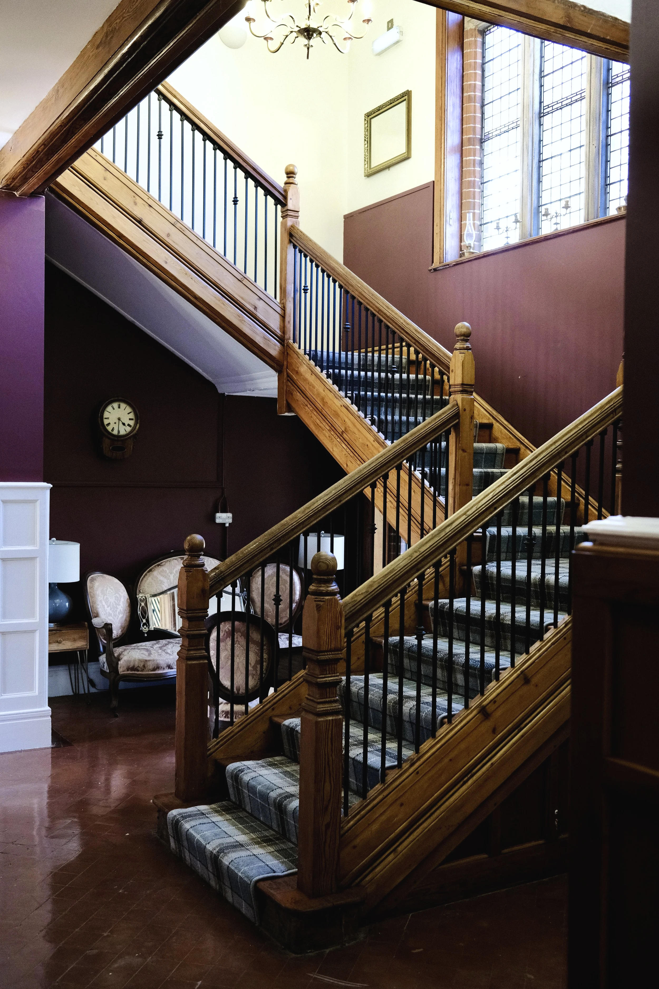 Interior view of a staircase with wooden railings and black iron balusters, carpeted steps, and a large stained glass window letting in natural light, in a decorated interior space with Victorian-style furniture and wall clock.
