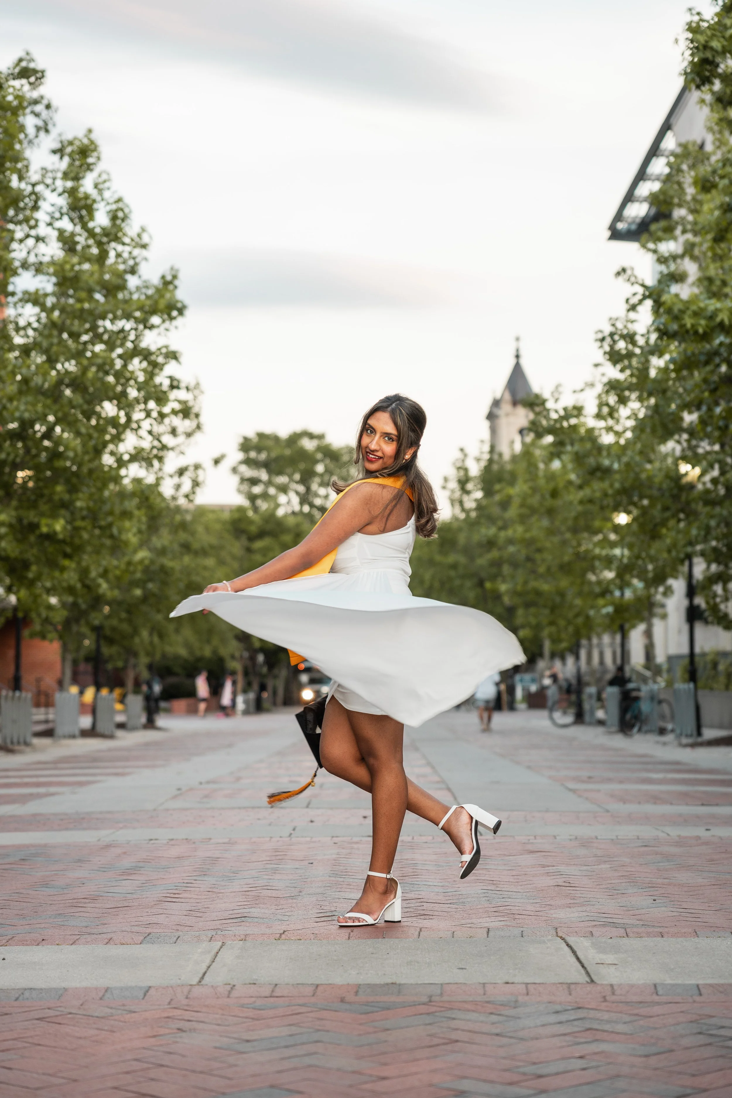 A woman in a white dress and high heels twirls on a city street with trees and buildings in the background.