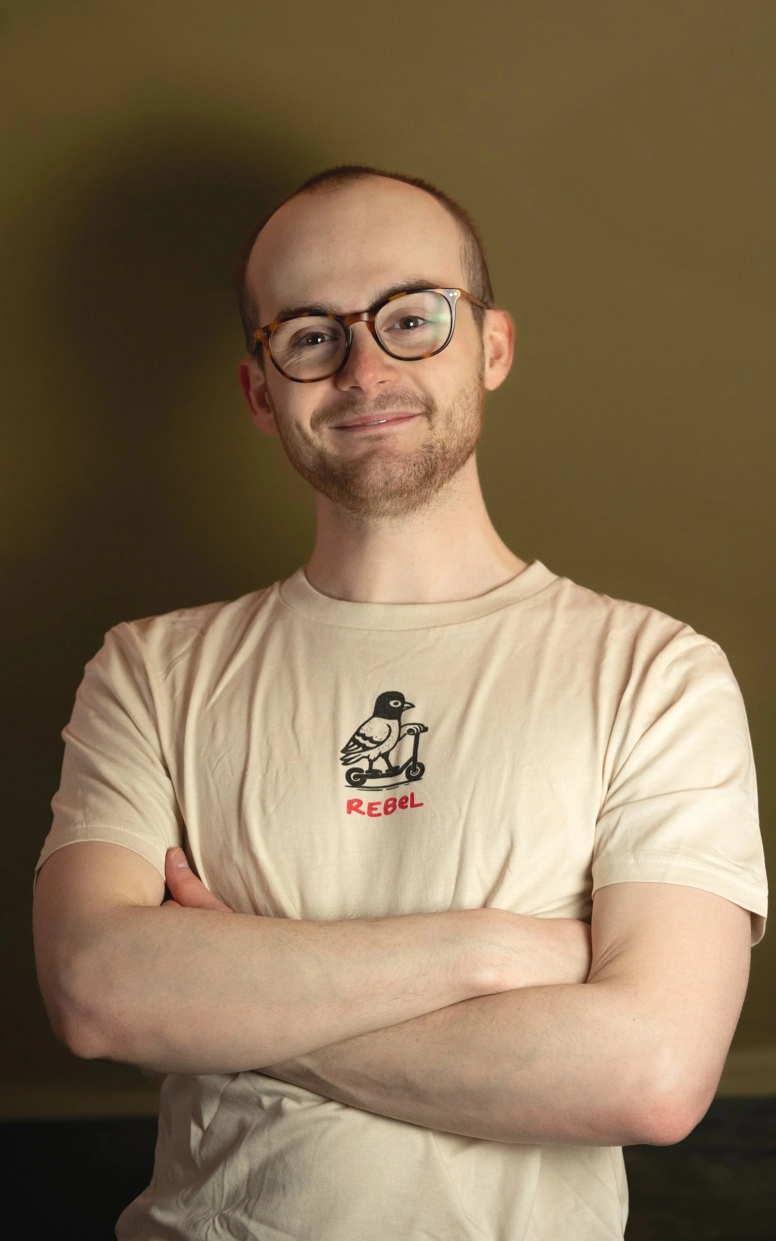A man with glasses, short hair, and a beard is standing with his arms crossed, smiling in front of a plain brown background. He is wearing a beige T-shirt with a bird graphic and the word 'REBEL' printed underneath.