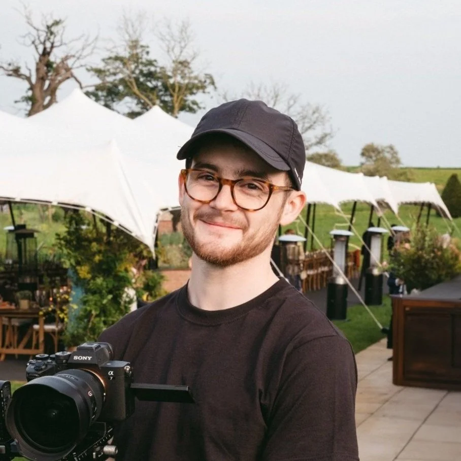 A young man with glasses and a black cap holds a camera outdoors at a venue with white canopy tents in the background.