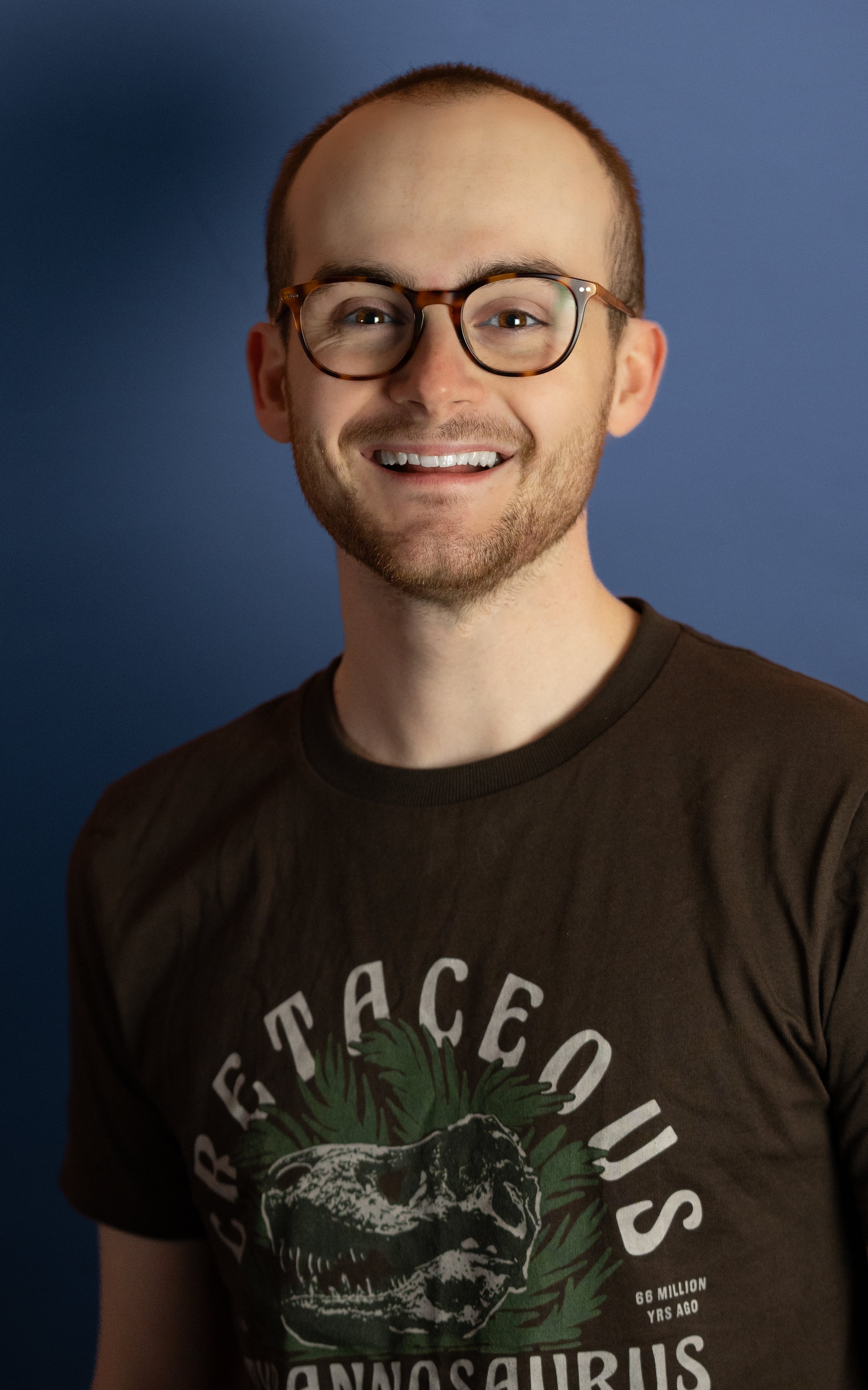 A young man with glasses smiling against a blue background, wearing a dark T-shirt with a dinosaur graphic and text.