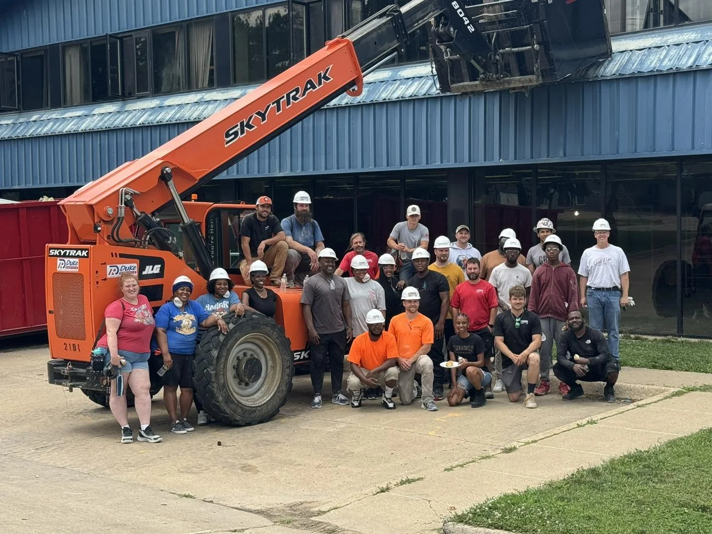 Group of diverse people with construction helmets gathered around a large orange skytrak construction vehicle in front of a building with glass windows.
