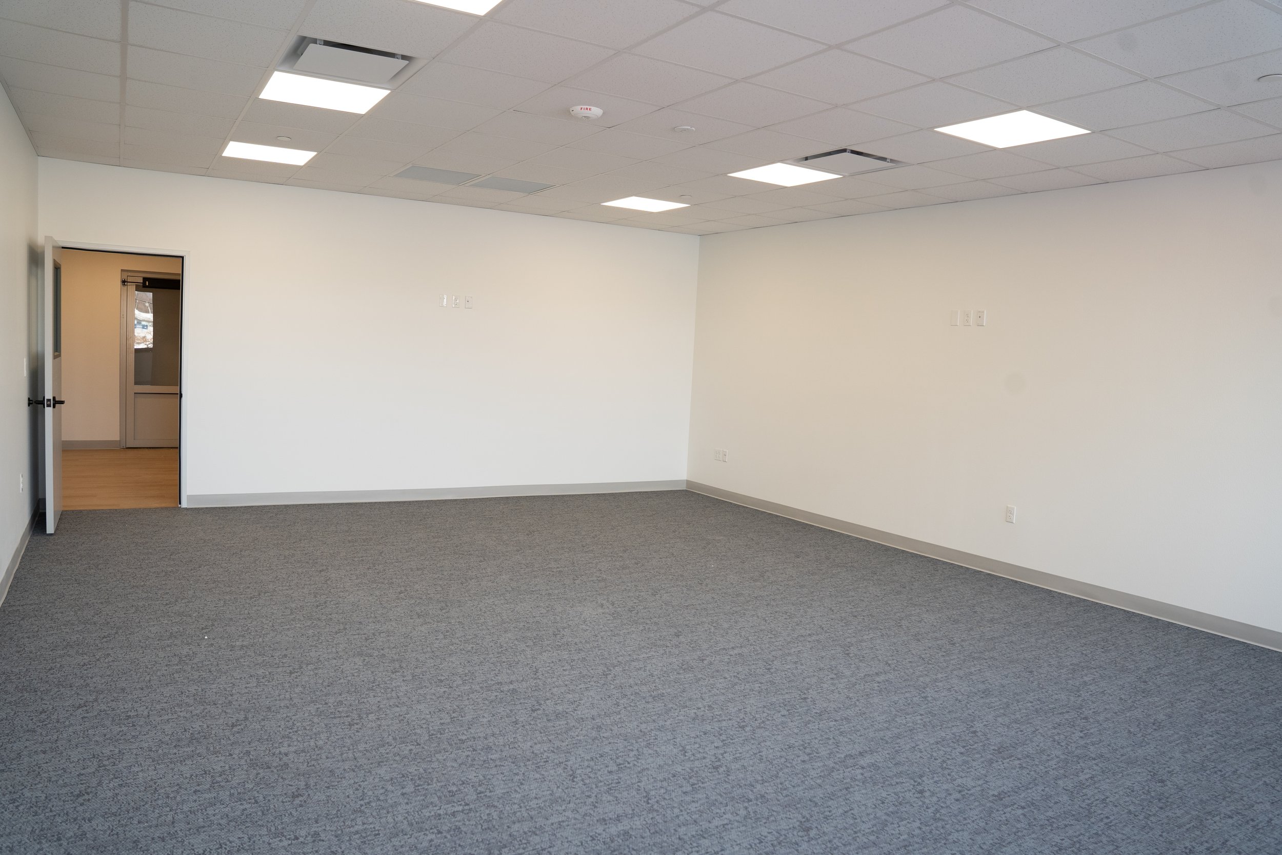 Empty office room with gray carpet, white walls, ceiling tiles, and a partially open door leading to another room.