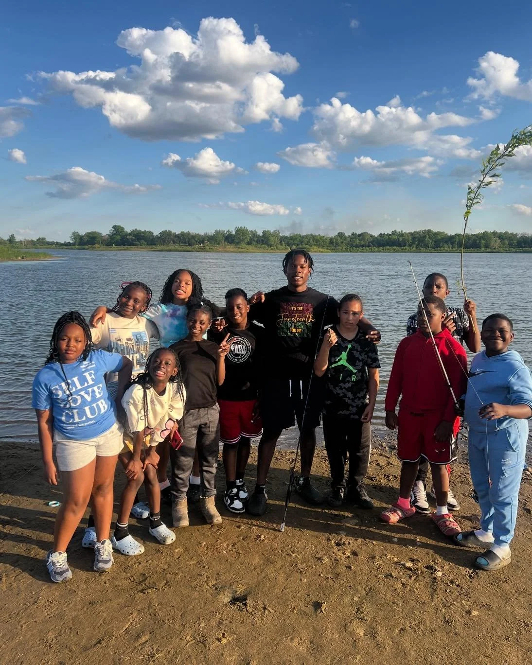 Group of children and a young man standing near a river on a sunny day, some holding sticks and one with a small tree branch, with a blue sky and clouds in the background.