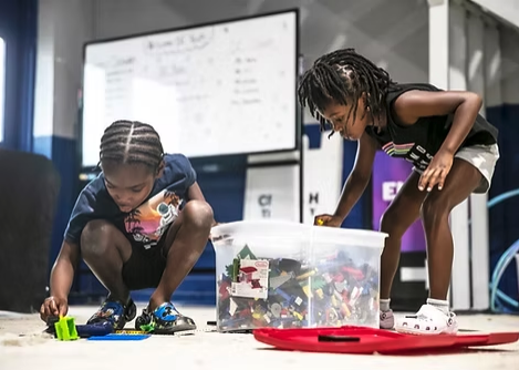 Two young girls playing with building blocks on the floor in front of a large whiteboard.