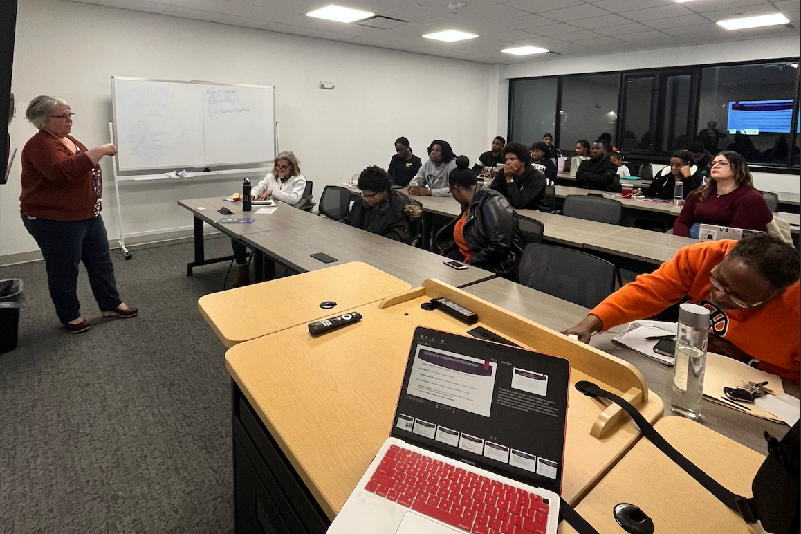 A classroom with a group of students sitting at desks and a woman standing at the front near a whiteboard, possibly giving a lecture.