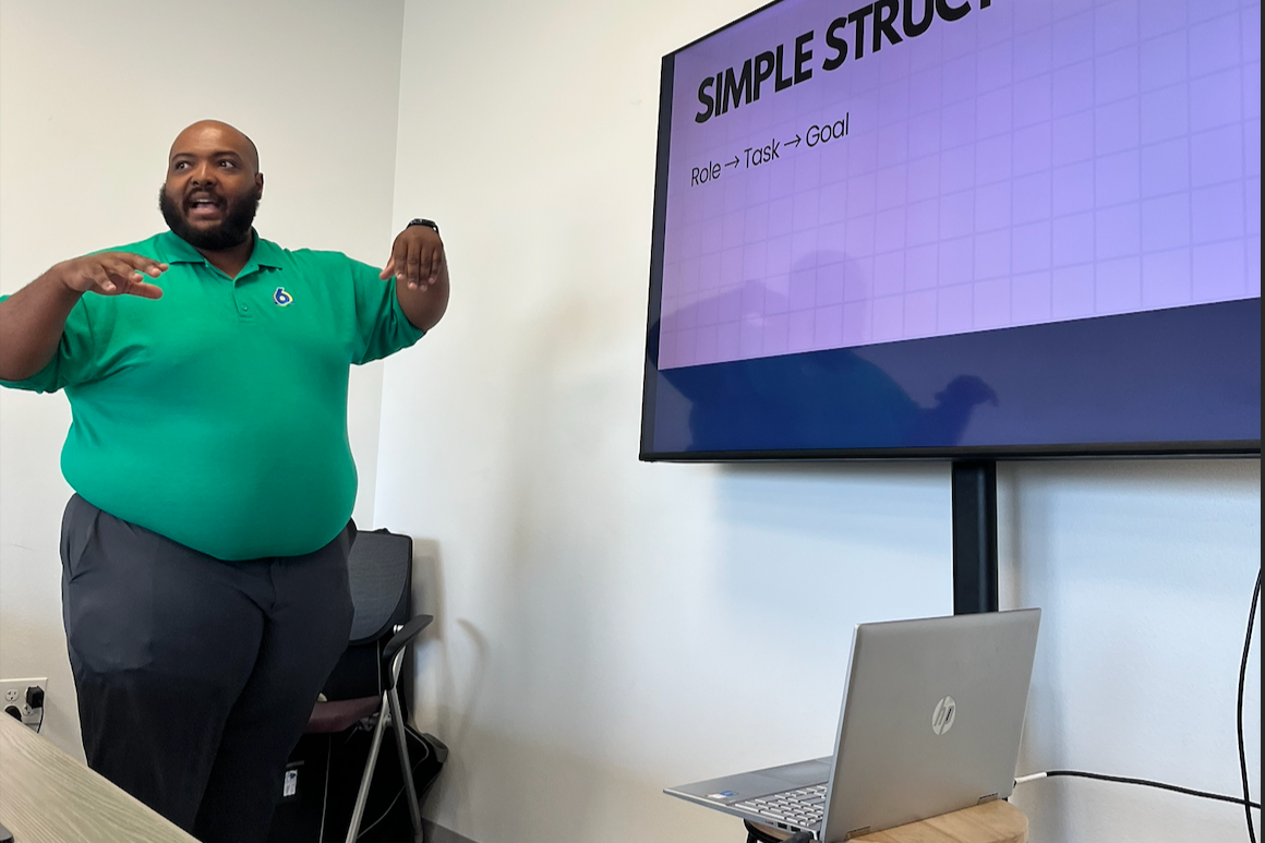 A man in a green polo shirt giving a presentation in a conference room with a large screen displaying a slide titled 'SIMPLE STRUCTURE' and the words 'Role -> Task -> Goal' underneath.