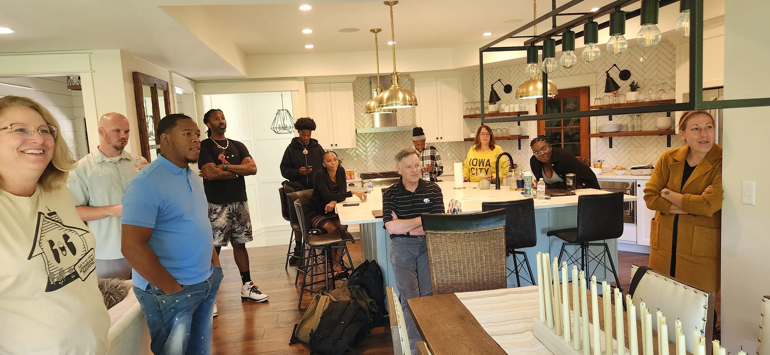 A group of people in a kitchen, some standing and some sitting, listening to a woman speaking. The kitchen is modern with white cabinets, pendant lights, and open shelves.
