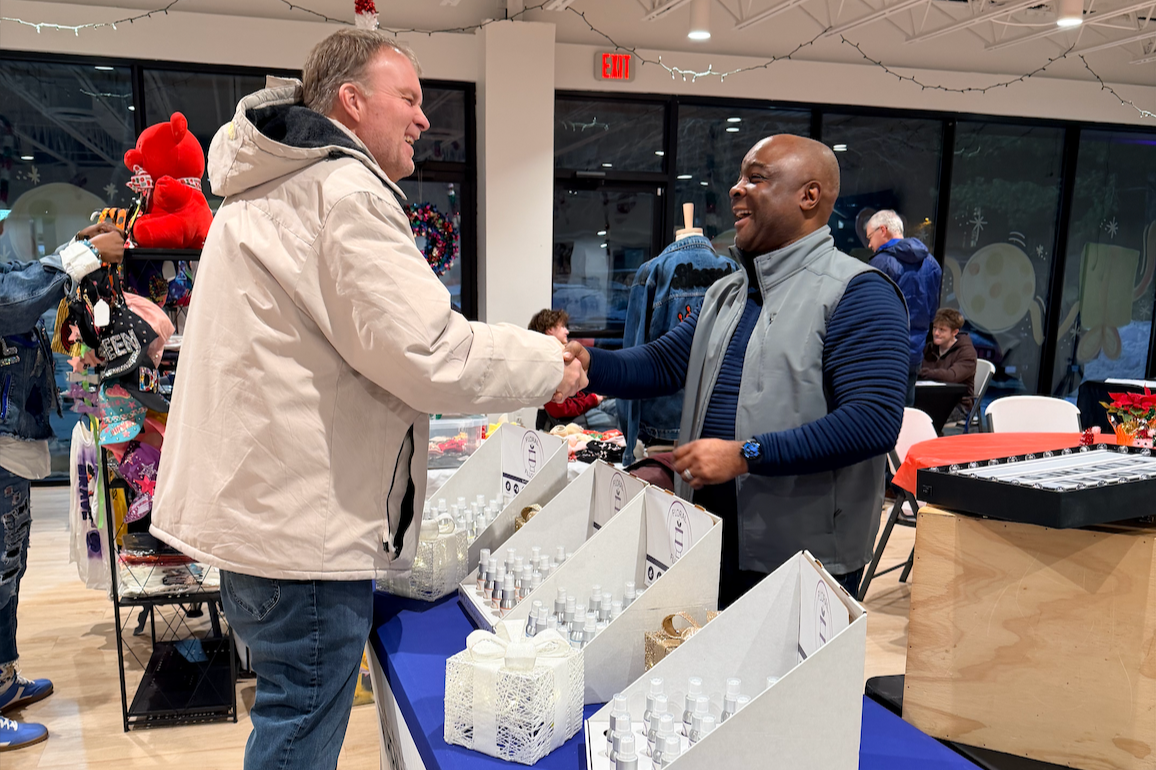 Two men shaking hands and smiling at a craft fair or market. There are tables with bottles and jewelry for sale. Holiday decorations are visible, including a Christmas wreath and red plush reindeer.