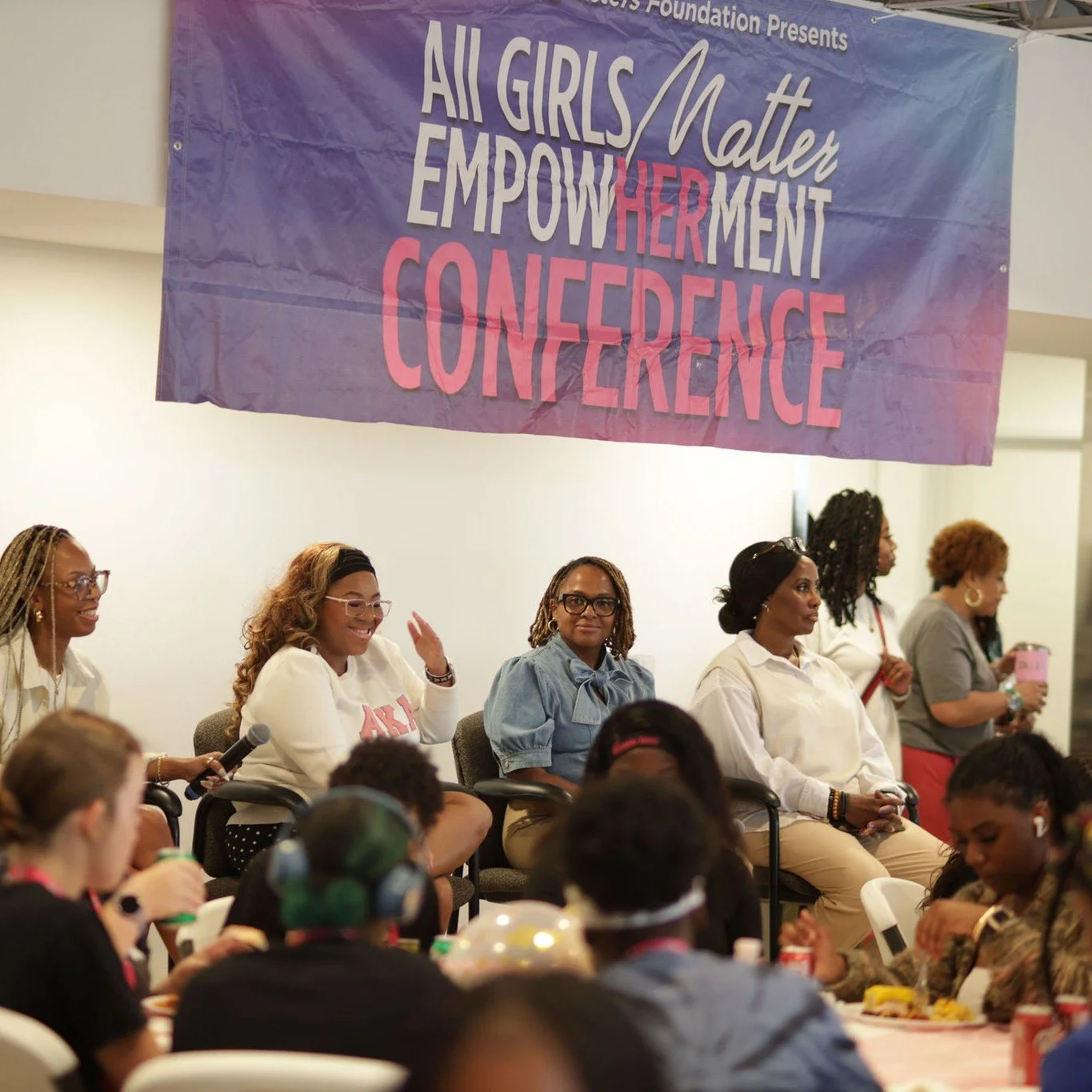 Women sitting on a stage, participating in a conference with a large purple banner behind them that reads 'All Girls Matter Empower Her Ment Conference.'