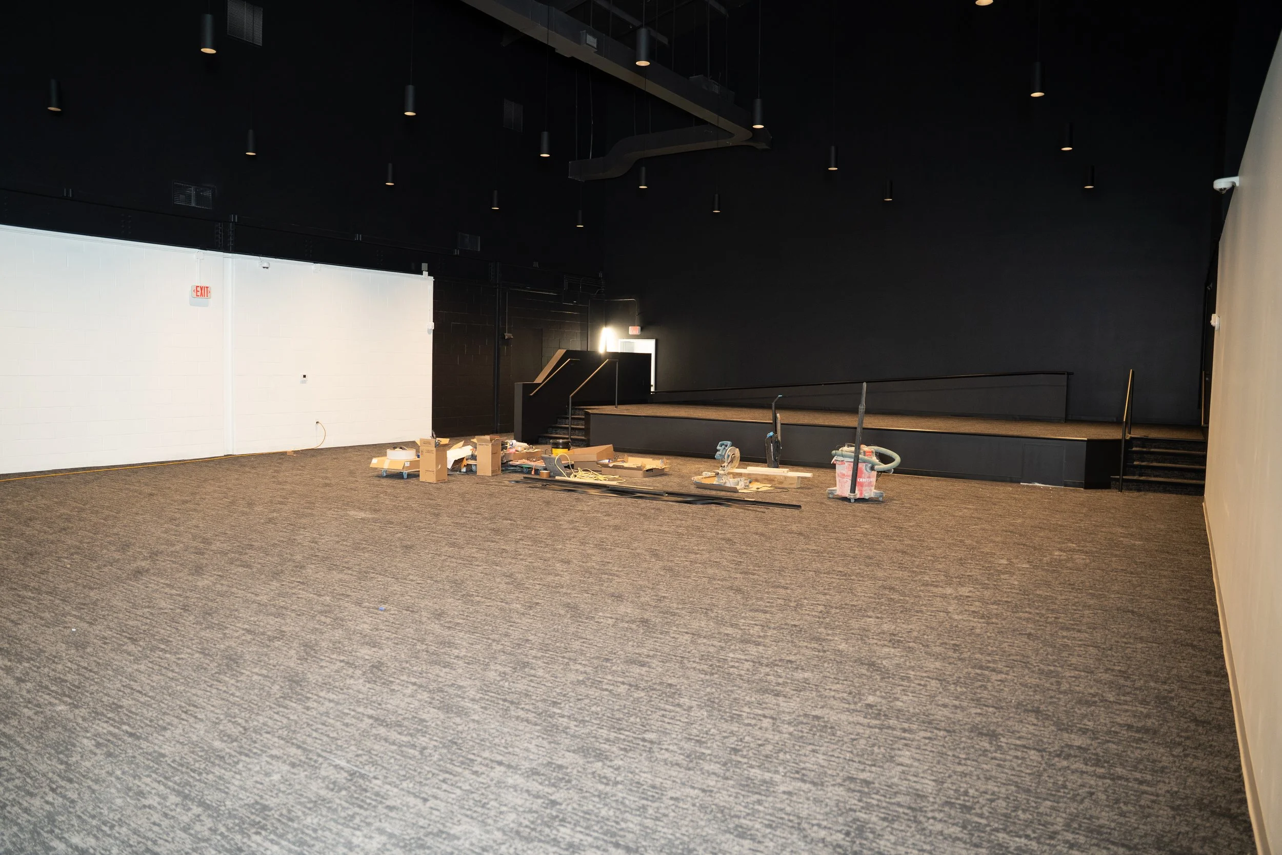 Empty room with dark walls, a small stage with stairs, and construction materials on the floor.