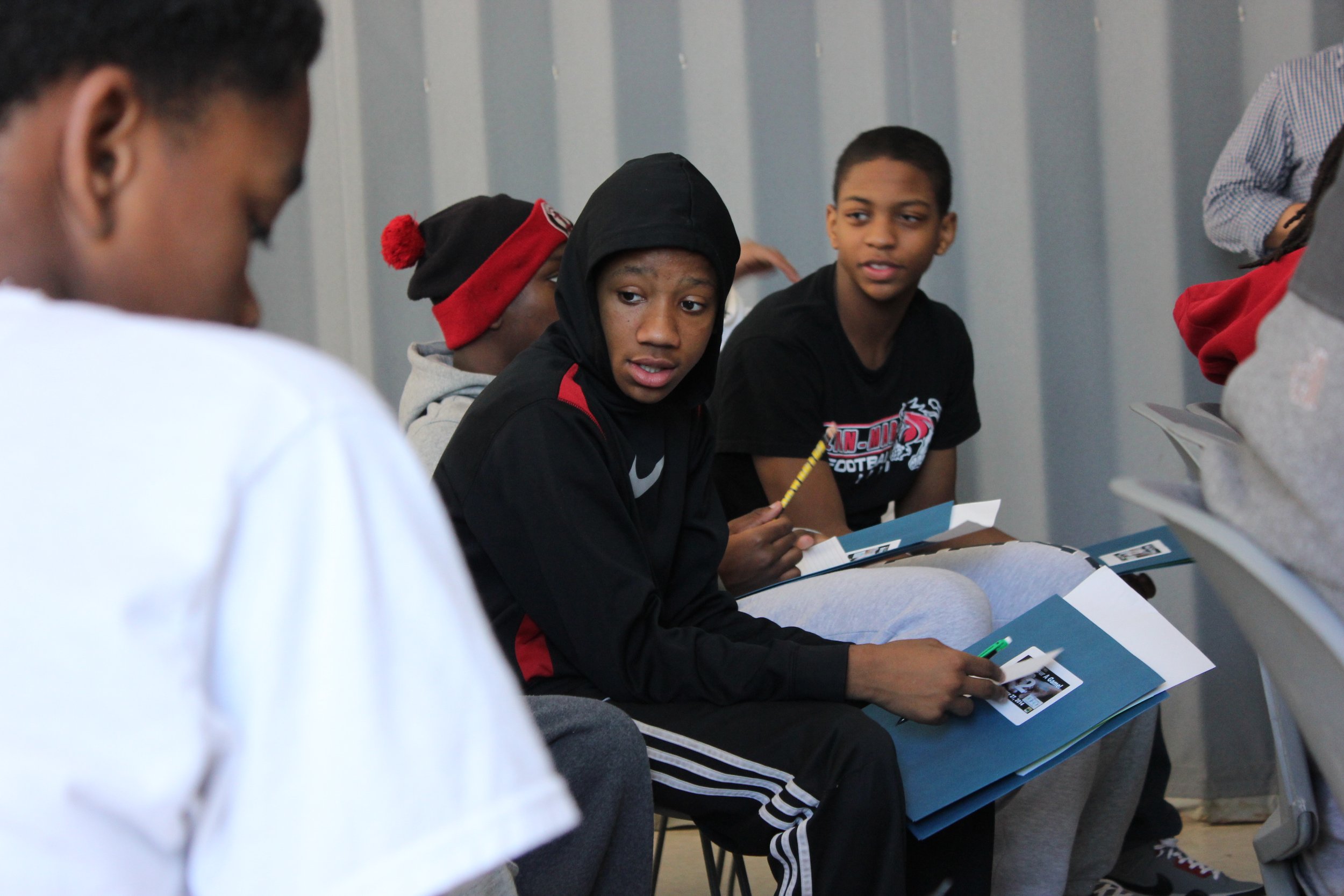 Group of young boys sitting in a line, talking and holding folders with photos inside, in a classroom or meeting space.