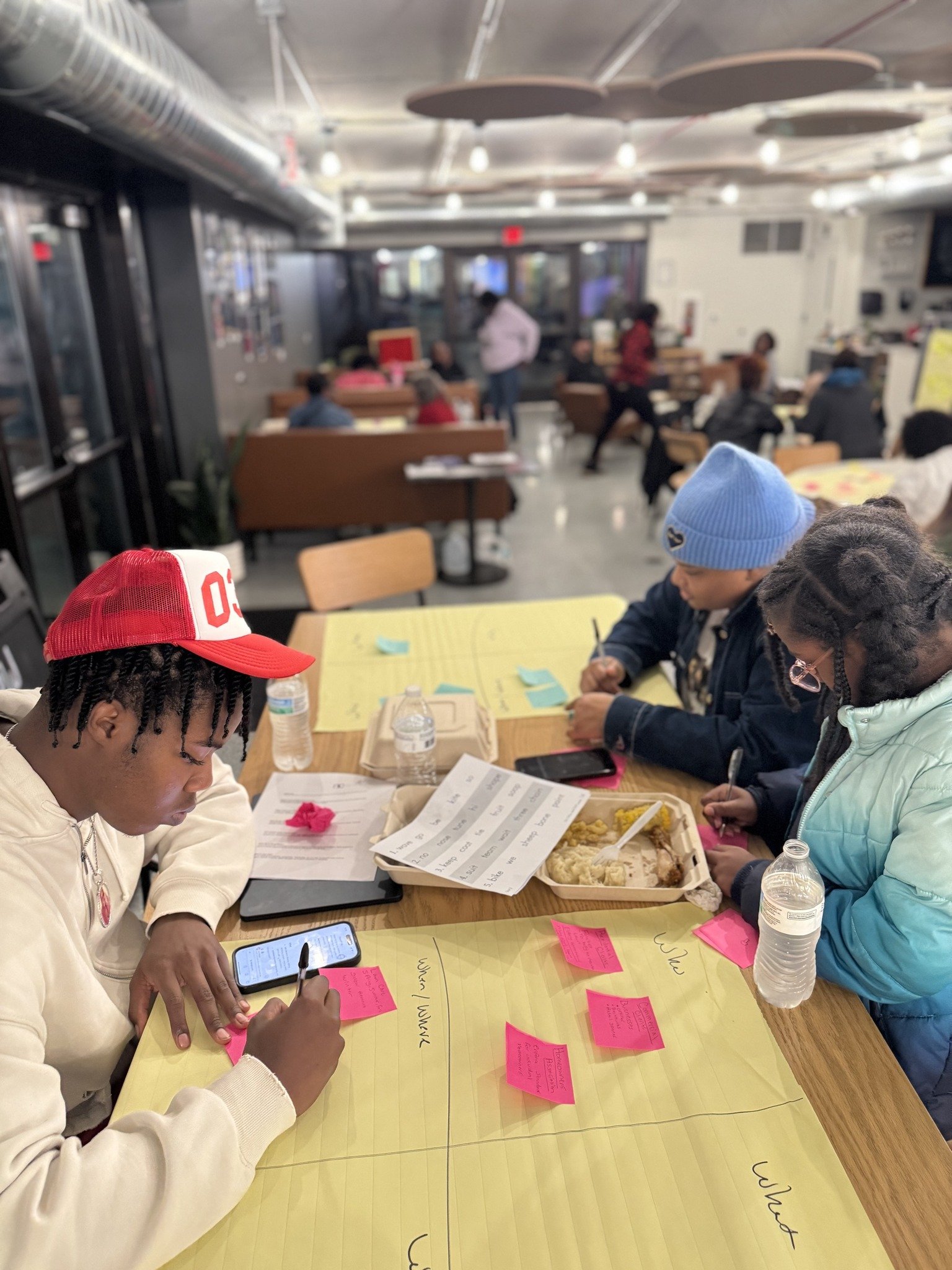 Three children sit at a table in a busy indoor setting, working on sticky notes and writing. One is wearing a red and white cap, another a blue beanie, and the third has glasses and is in a light-colored jacket. There are water bottles, a food container, and papers on the table, with people and activity in the background.