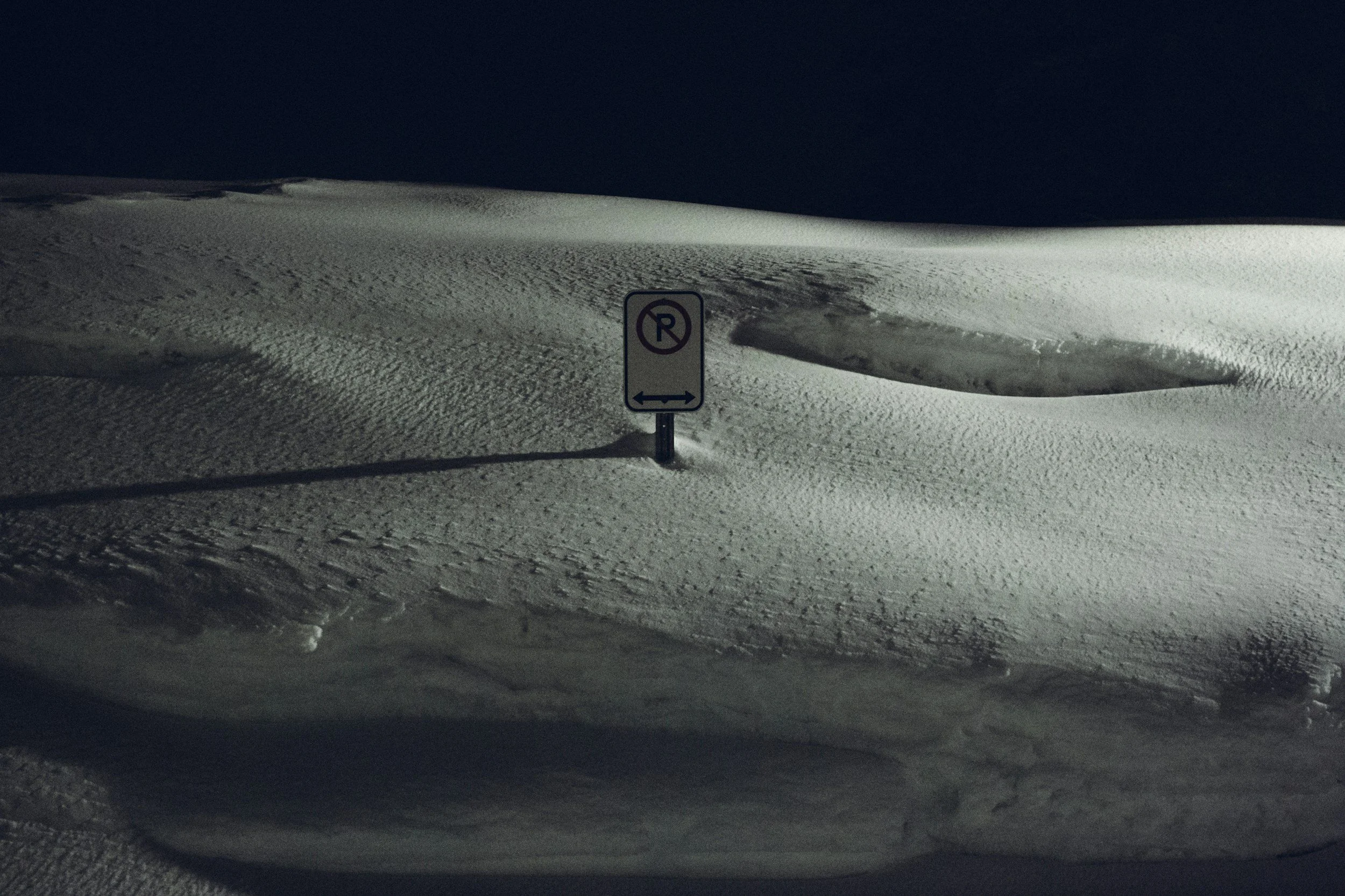 Snow-covered landscape with a 'No Parking' sign and no visible vehicles, illuminated by artificial light at night.