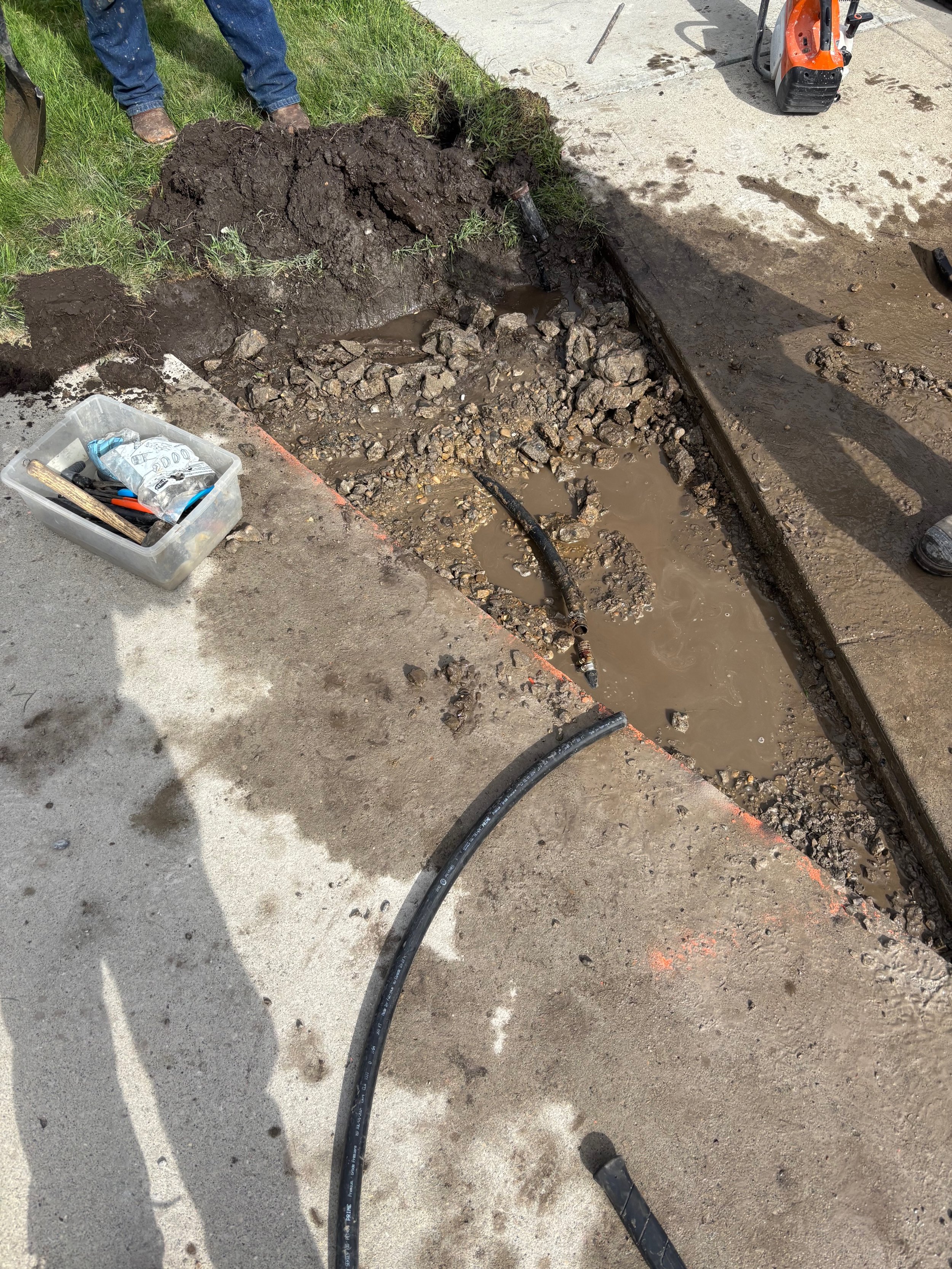 A construction site showing an excavated trench with muddy water, black hoses, and dirt. Construction tools and equipment are visible, and two workers are present, with only their legs visible.
