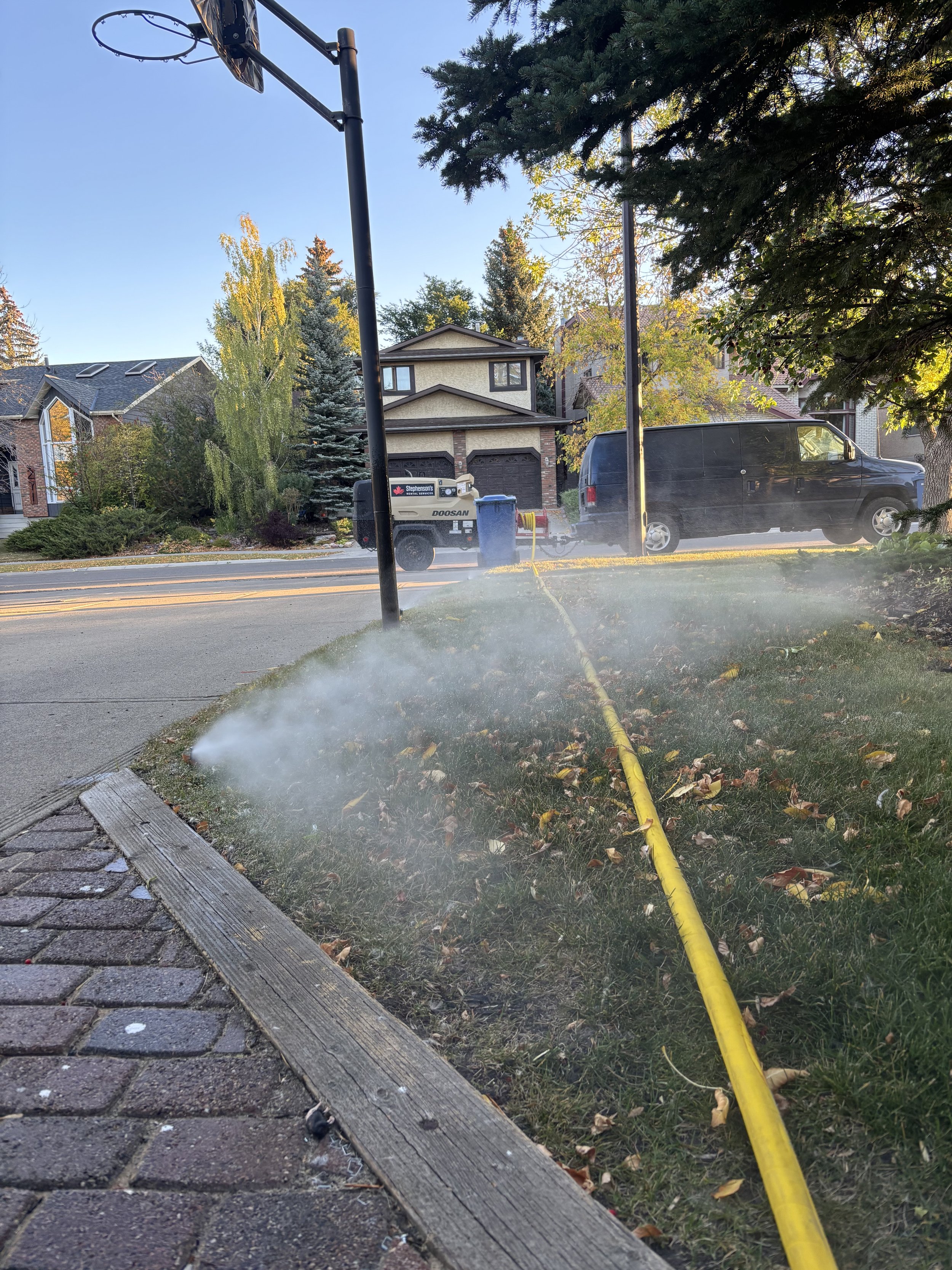 A hose connected to a yellow pipe sprays water onto the grass next to a sidewalk and street in a residential neighborhood with houses and trees in the background.