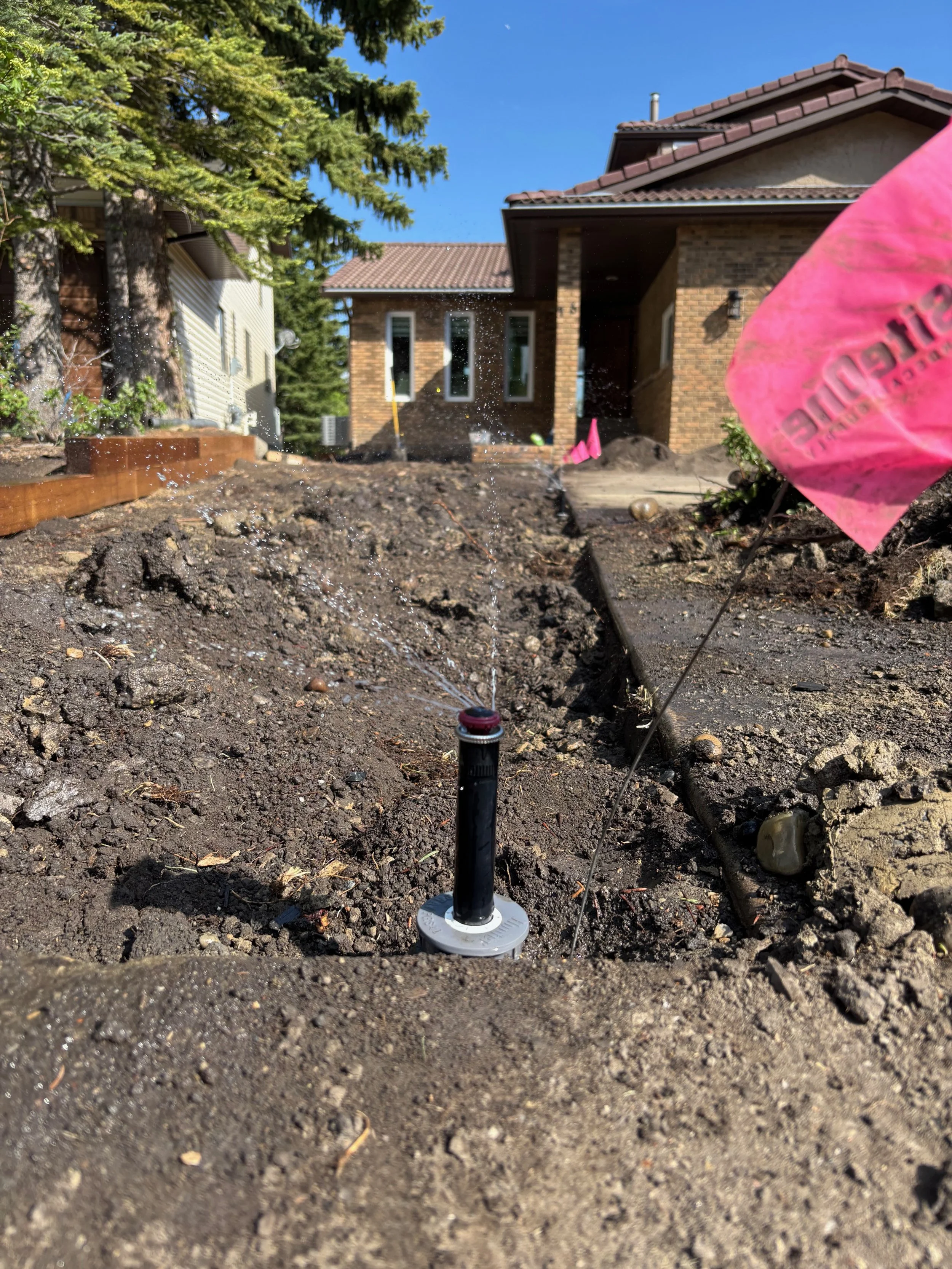A soil site with a sprinkler watering the dirt in a residential backyard, with a brick house, trees, and a blue sky in the background.
