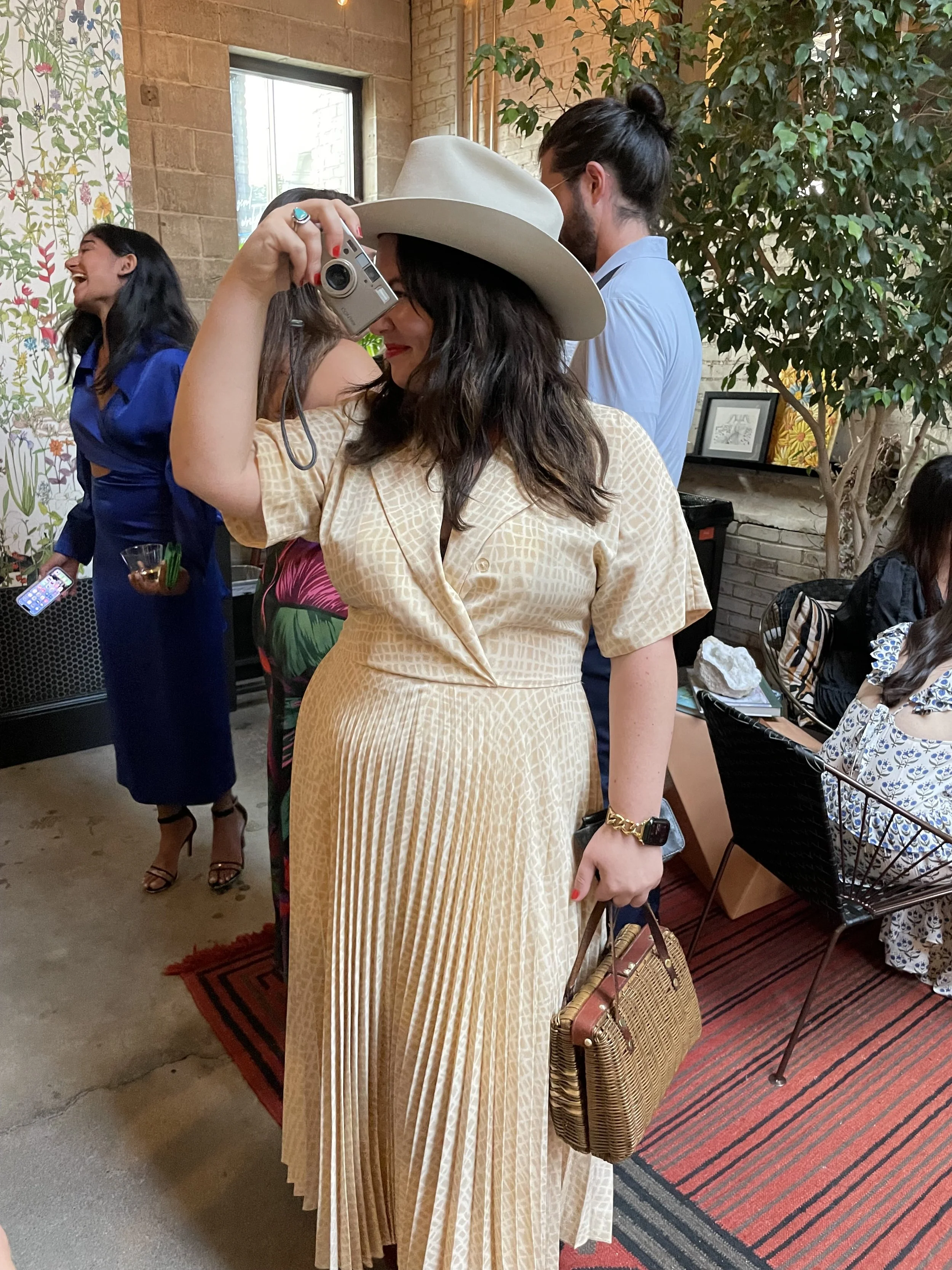 Woman in a beige dress and white hat taking a photo with a point-and-shoot camera in a stylish indoor setting.