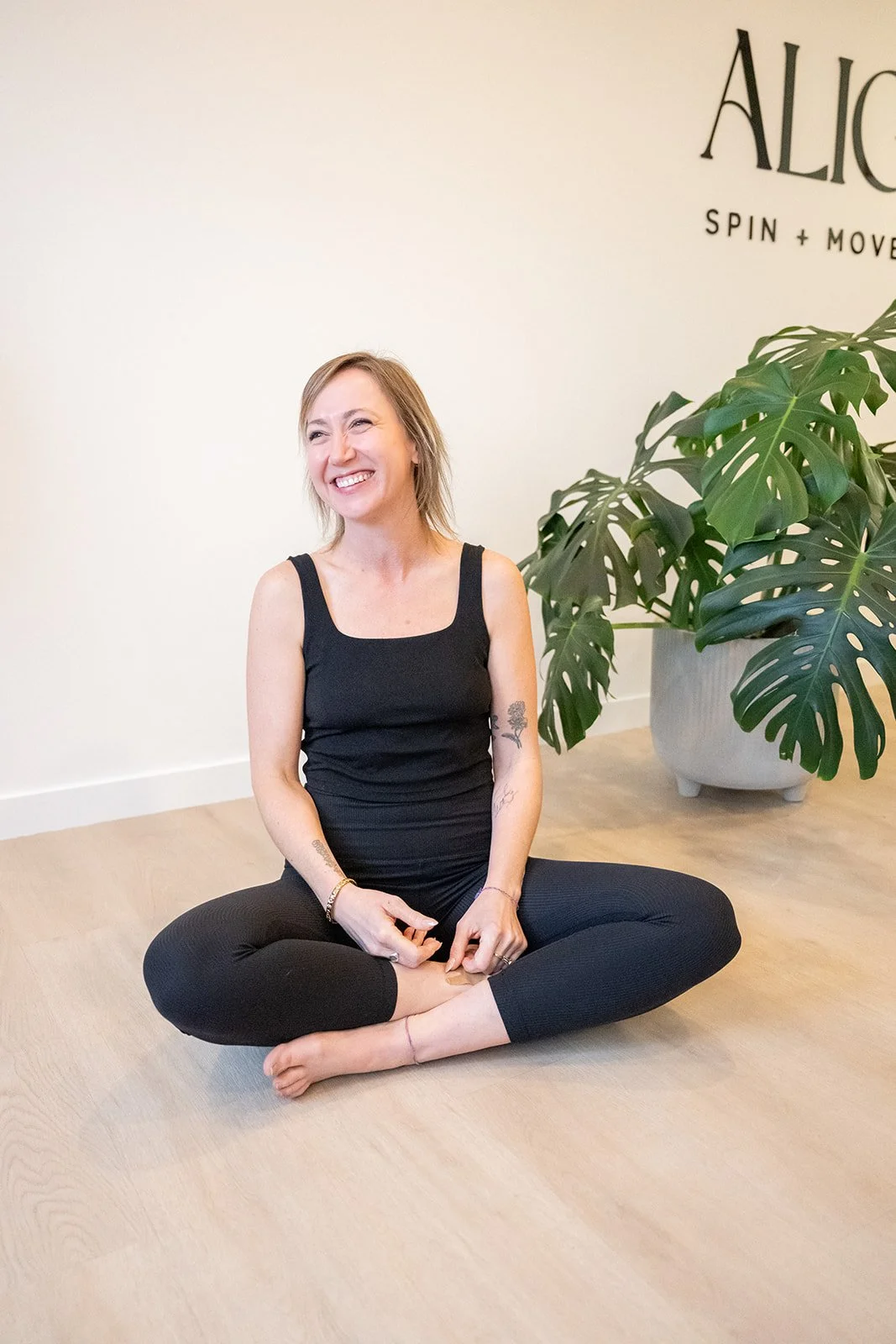 A woman with long red hair smiling and sitting cross-legged on the floor in front of large green houseplants, dressed in a black tank top and black leggings.