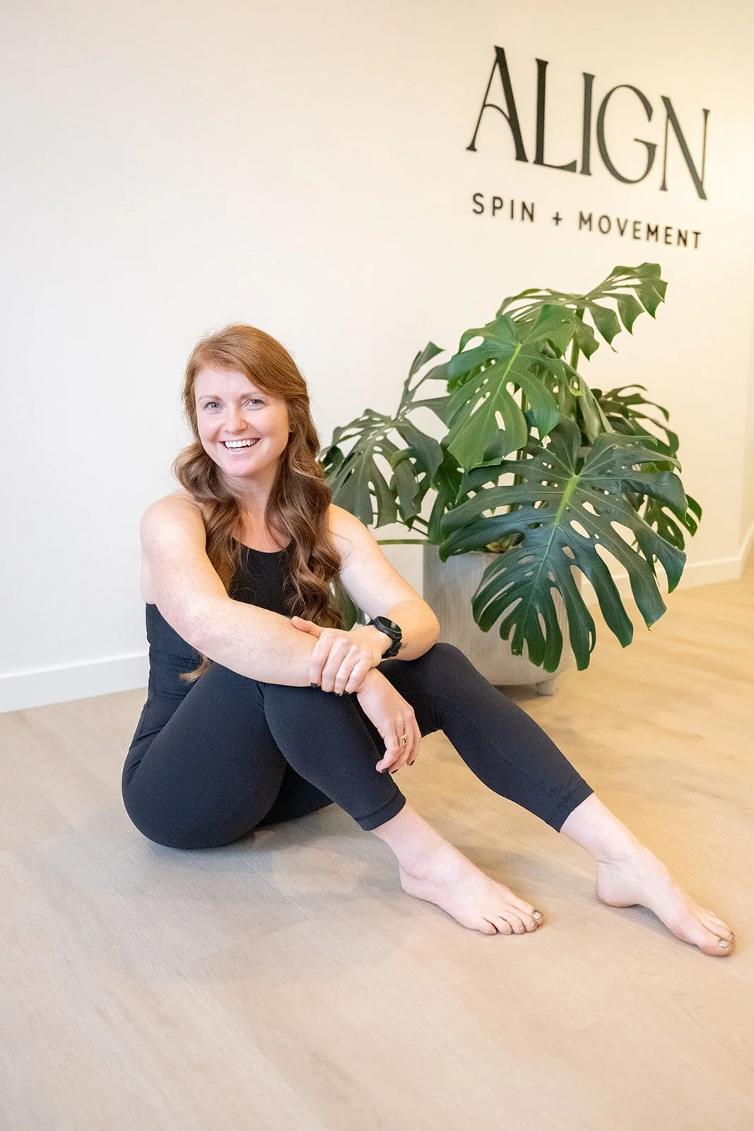 A woman with long red hair sitting on a wooden floor, smiling, wearing a black long-sleeve top, light blue shorts, and white socks with black stripes, in front of large houseplants in white pots.