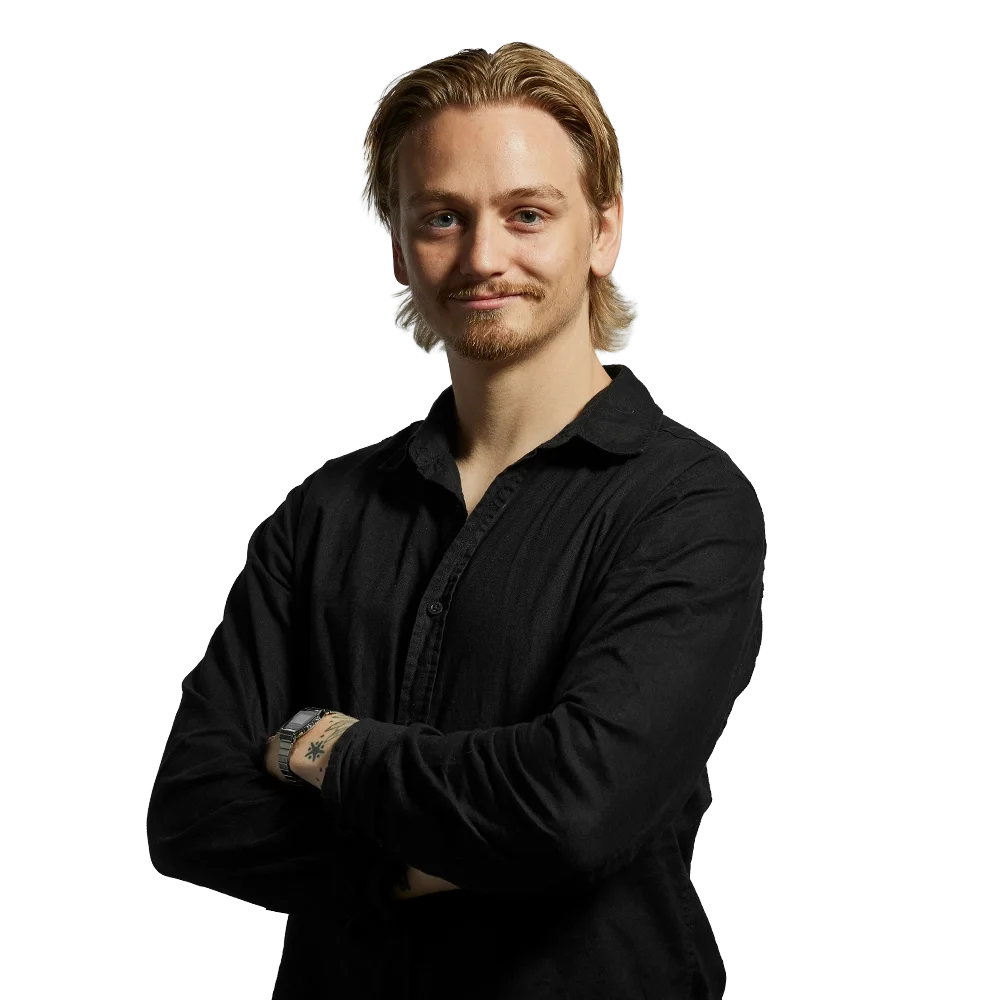 En ung man med blont hår, skägg, i svart skjorta, står med armarna korsade och ler.