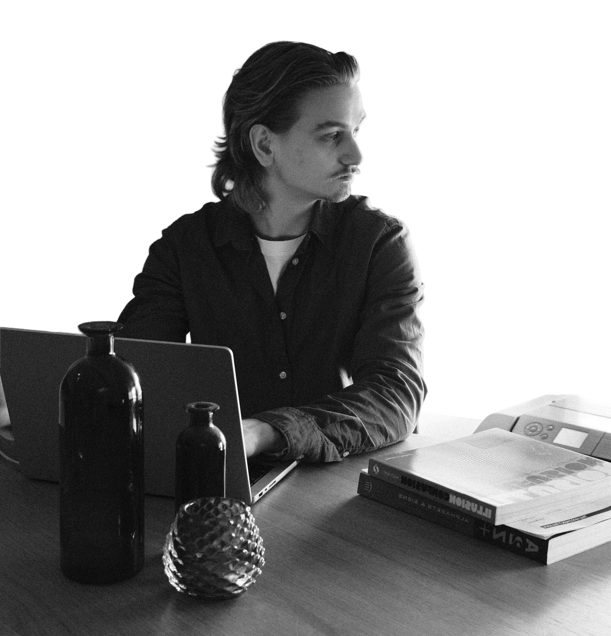 En man med långt hår sitter vid ett skrivbord, tittar åt sidan. På bordet finns en laptop, några böcker, en pärm, och några vaser.