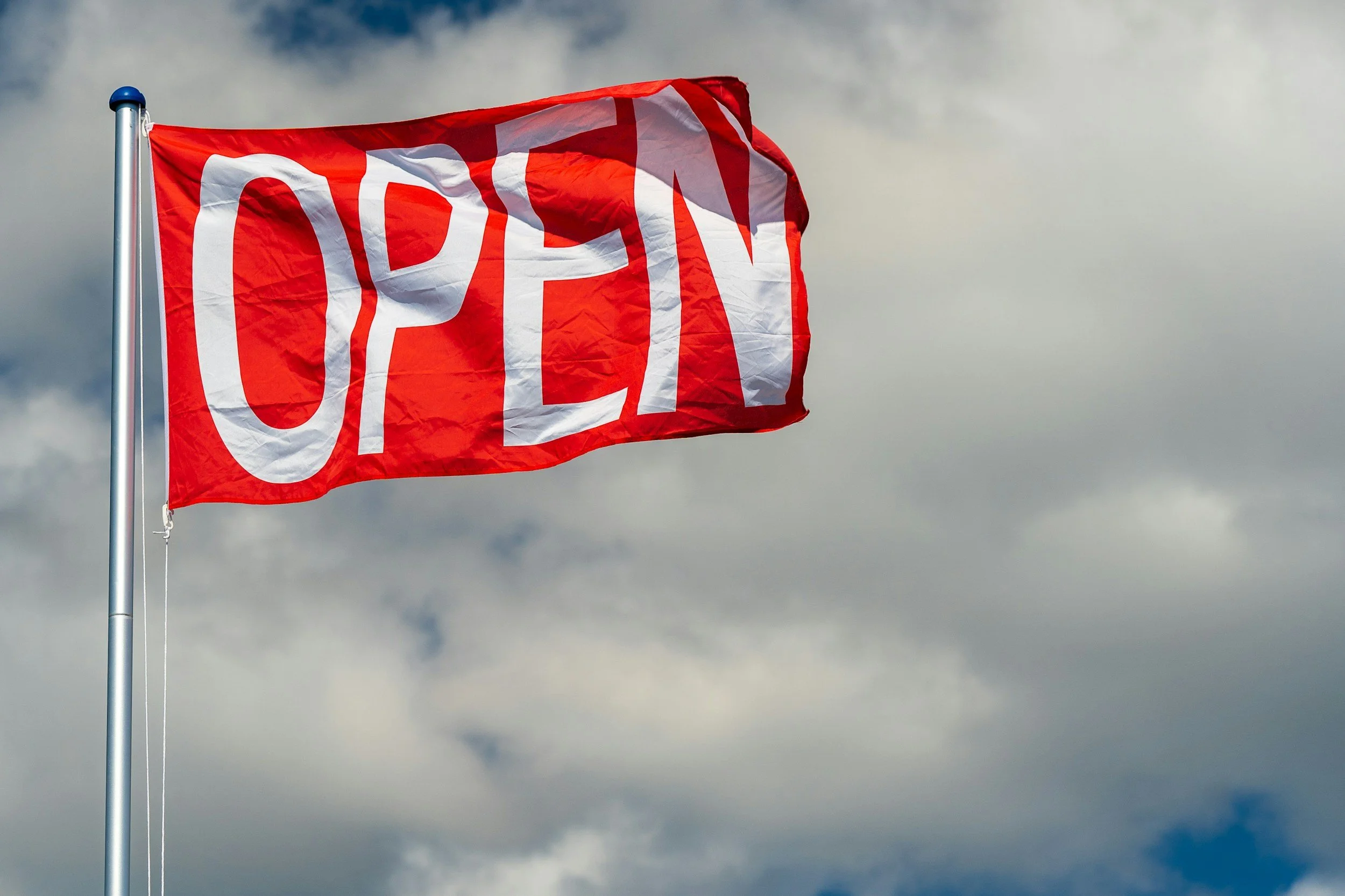 En röd flagga med vit text som säger "OPEN" vajar i vinden mot en molnig himmel.
