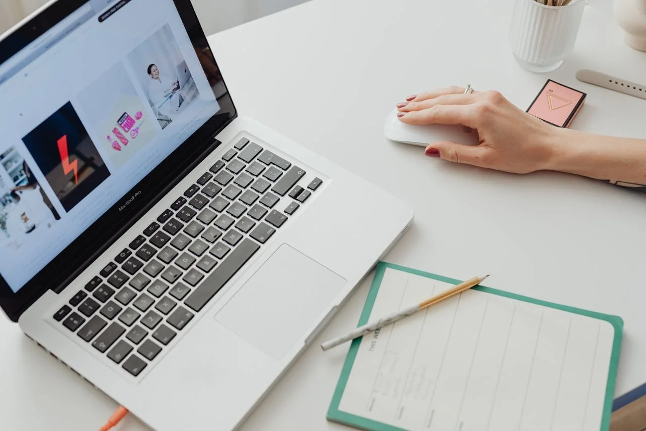 En hand som använder en trådlös mus på ett skrivbord. På skrivbordet finns en öppen laptop, en anteckningsbok och några andra föremål.