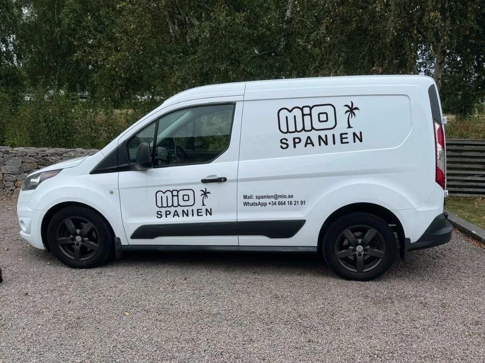 En vit leveranskasse med text och en palmpalm på sidan, står parkerad på en grusplan med träd i bakgrunden.