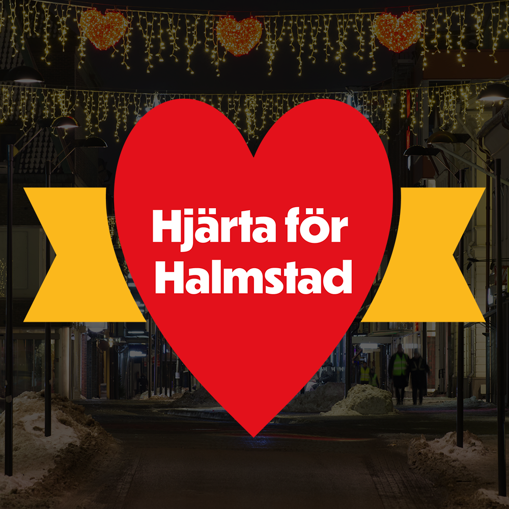 En stor röd hjärta med texten 'Hjärta för Halmstad' i vitt, omgivet av två gult band på vardera sida, hängande över en stadsgata dekorerad med julutskärningar och ljusslingor på kvällen.
