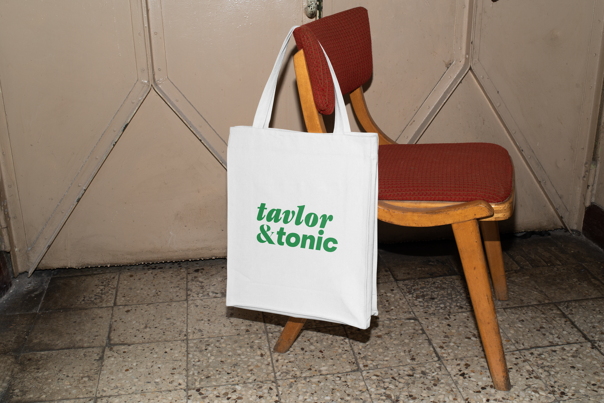 En vit tygväska med texten "tavlor & tonic" i grönt står lutad mot en vintage trästol med röd klädsel, placerad i ett rum med klinker golv och beige väggar.