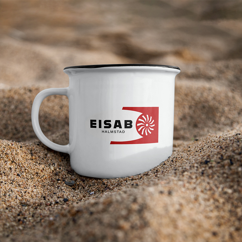 En vit mugg med svart insida och en logo för EISAB Halmstad, placerad i sanden på stranden.