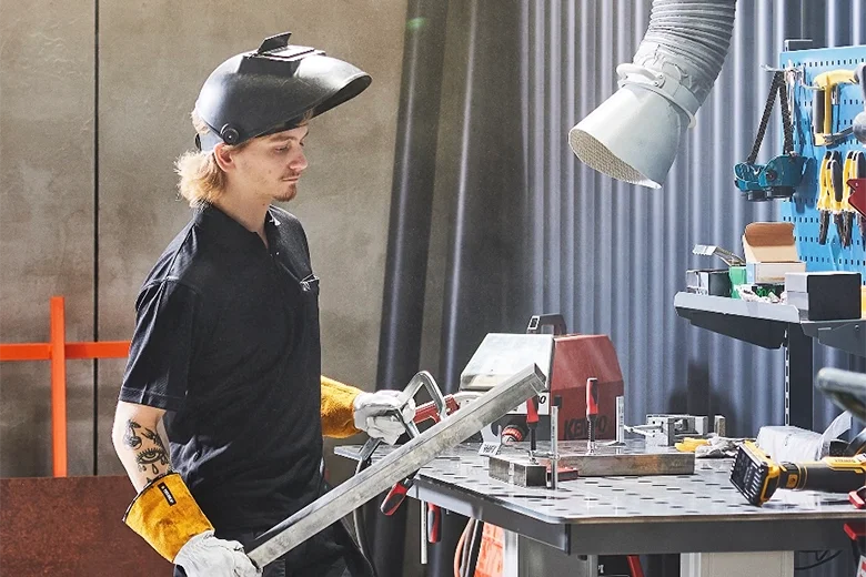 En person i skyddsglasögon och arbetskläder arbetar i ett verkstadslabb med verktyg och maskiner.