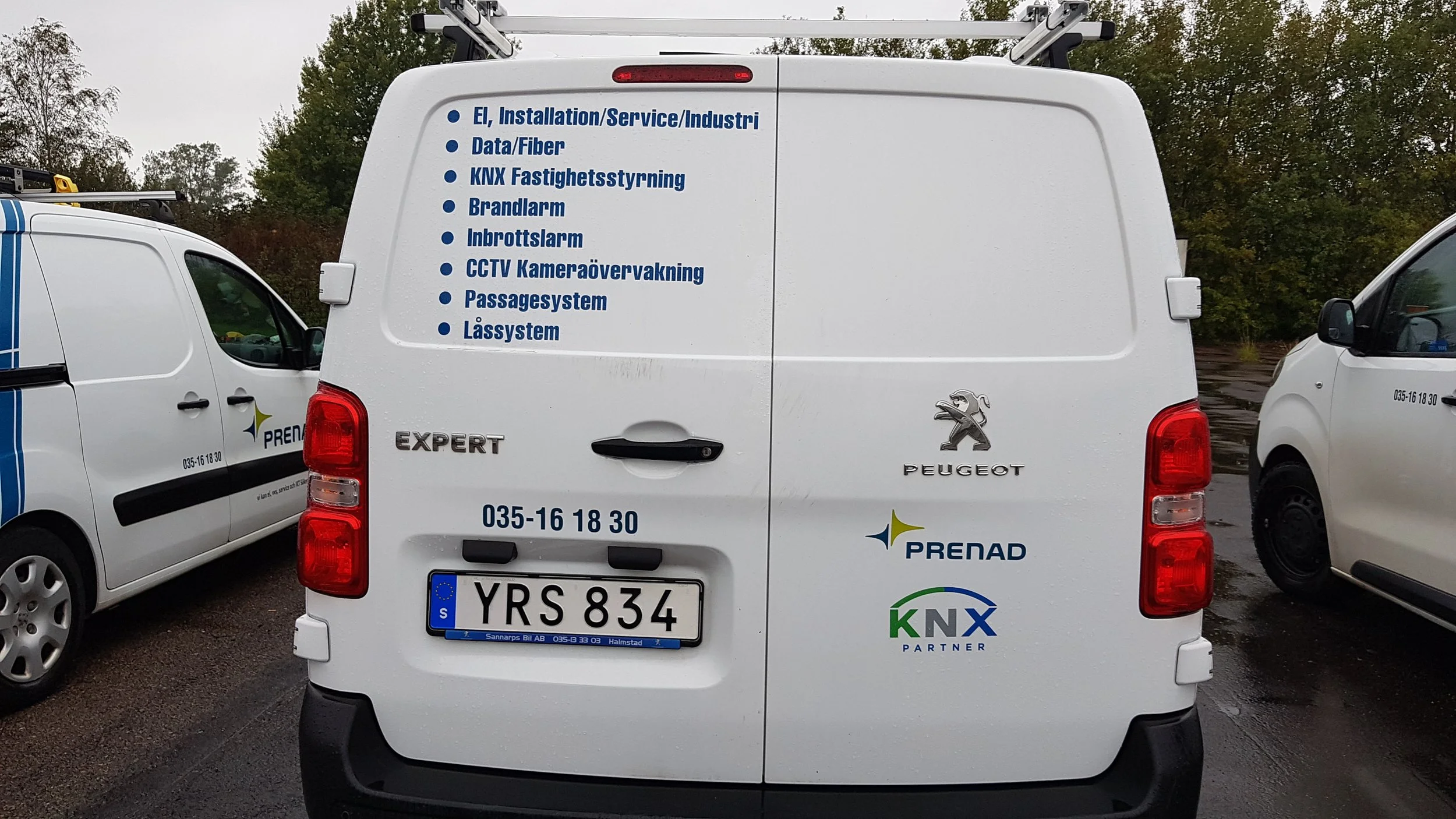 En vit Peugeot Expert-förvaringsbil med företagsnamn och tjänster listade på bakdörren, inklusive elinstallation, datafiber, larm, passage- och låssystem, på en parkeringsplats med andra vita bilar och träd i bakgrunden.